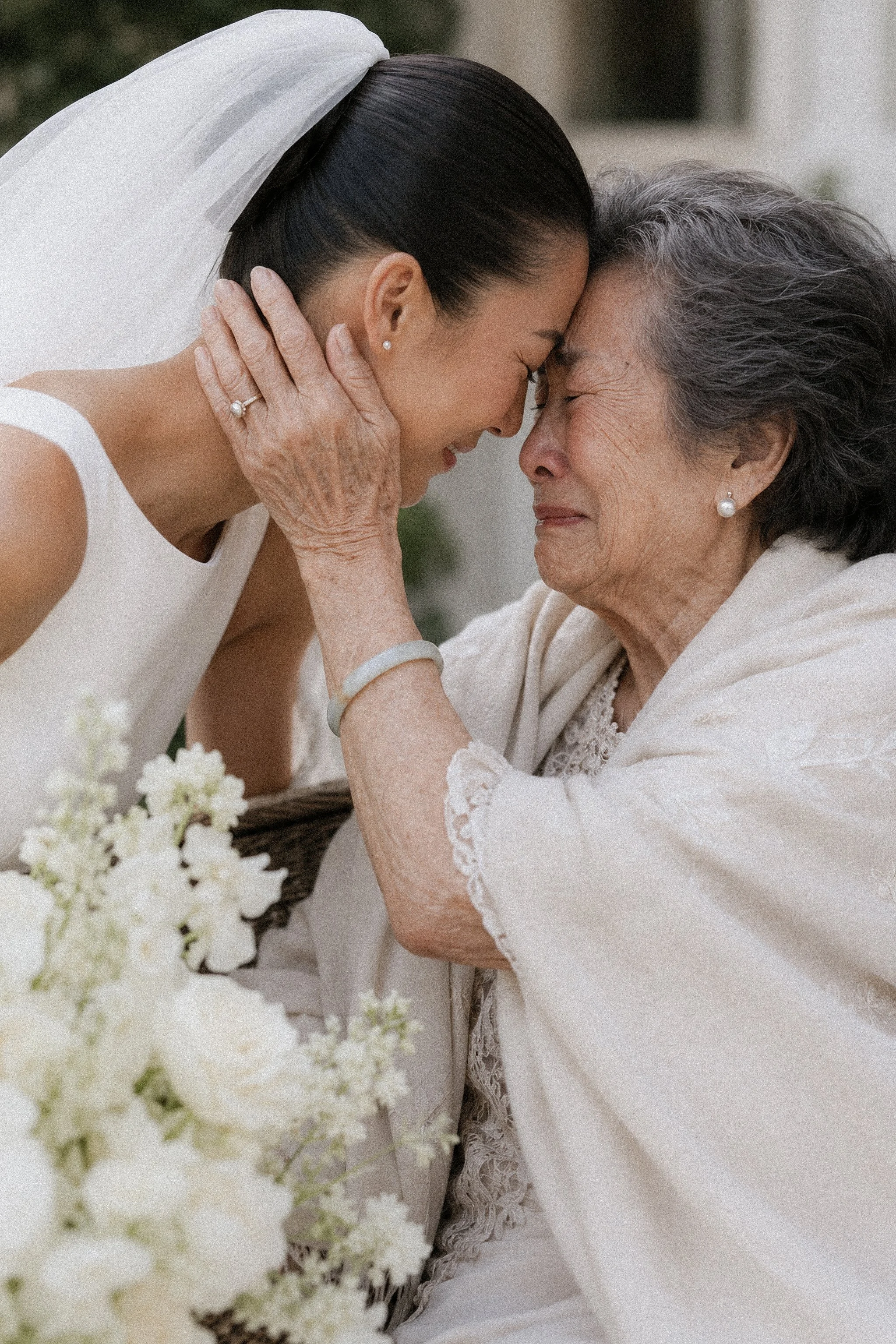 A tender moment between a bride and an elderly woman, possibly her grandmother, touching foreheads with their eyes closed and smiling, in a wedding or special event setting.