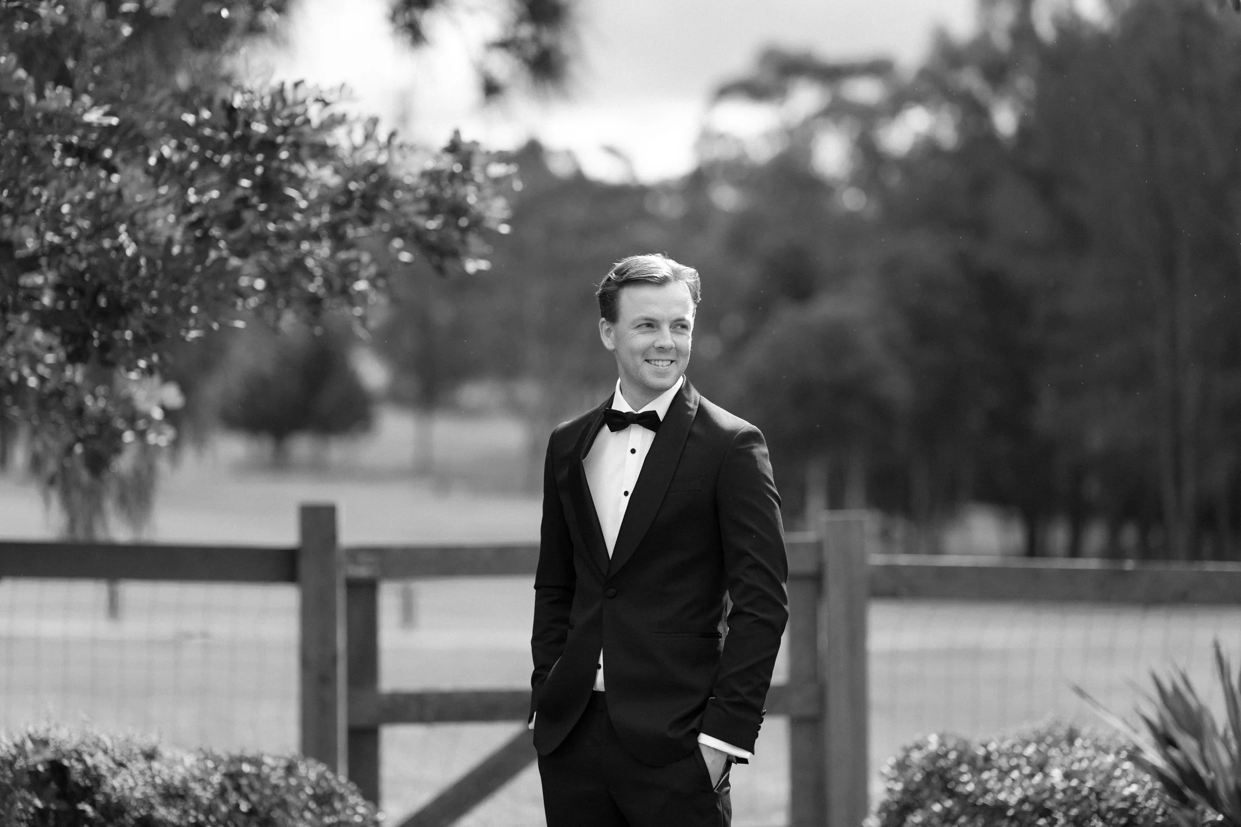 A man in a tuxedo with a bow tie standing outdoors near a fence and trees, smiling.