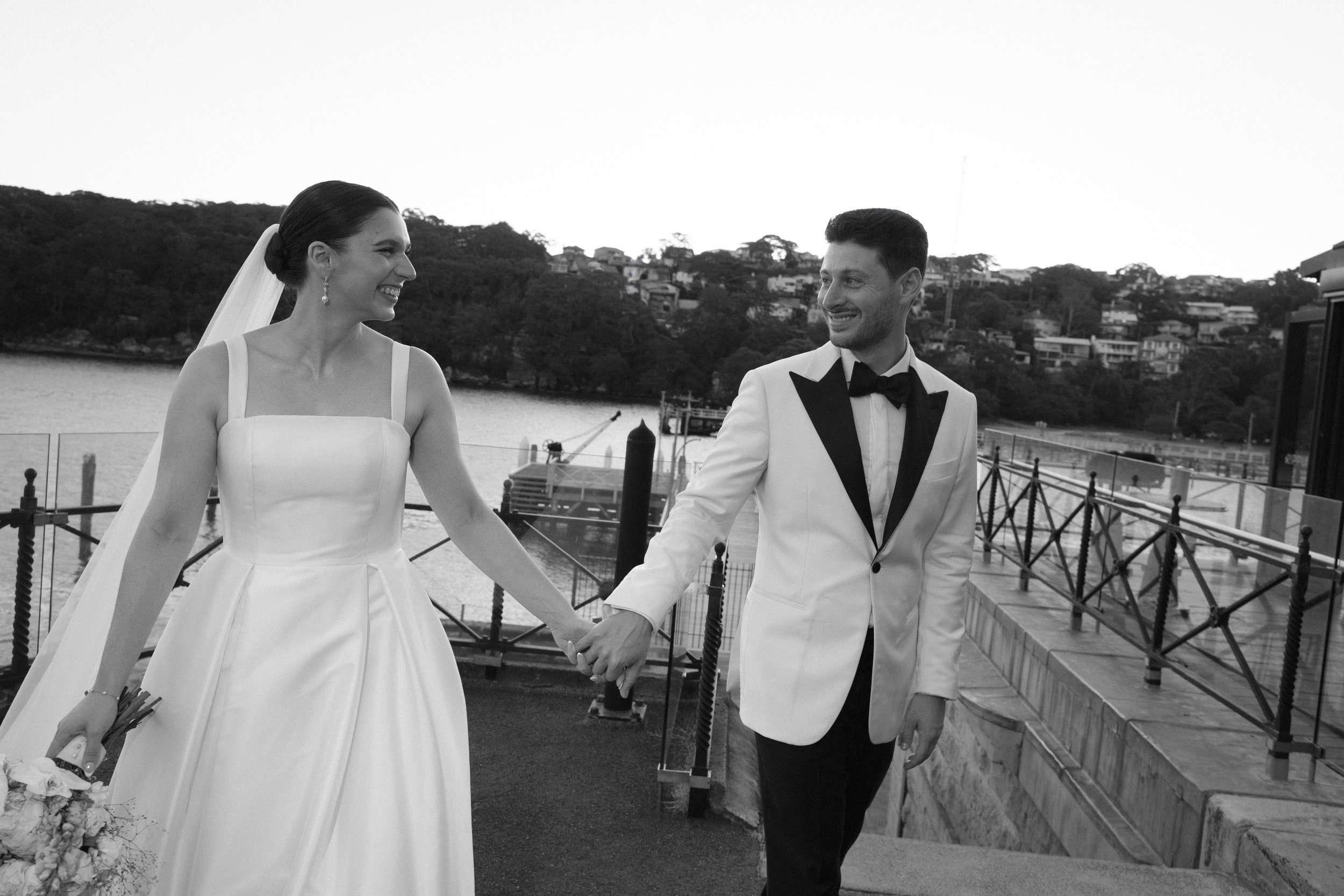 Black and white photo of a newlywed couple holding hands and smiling while walking along a waterfront with boats and hills in the background.
