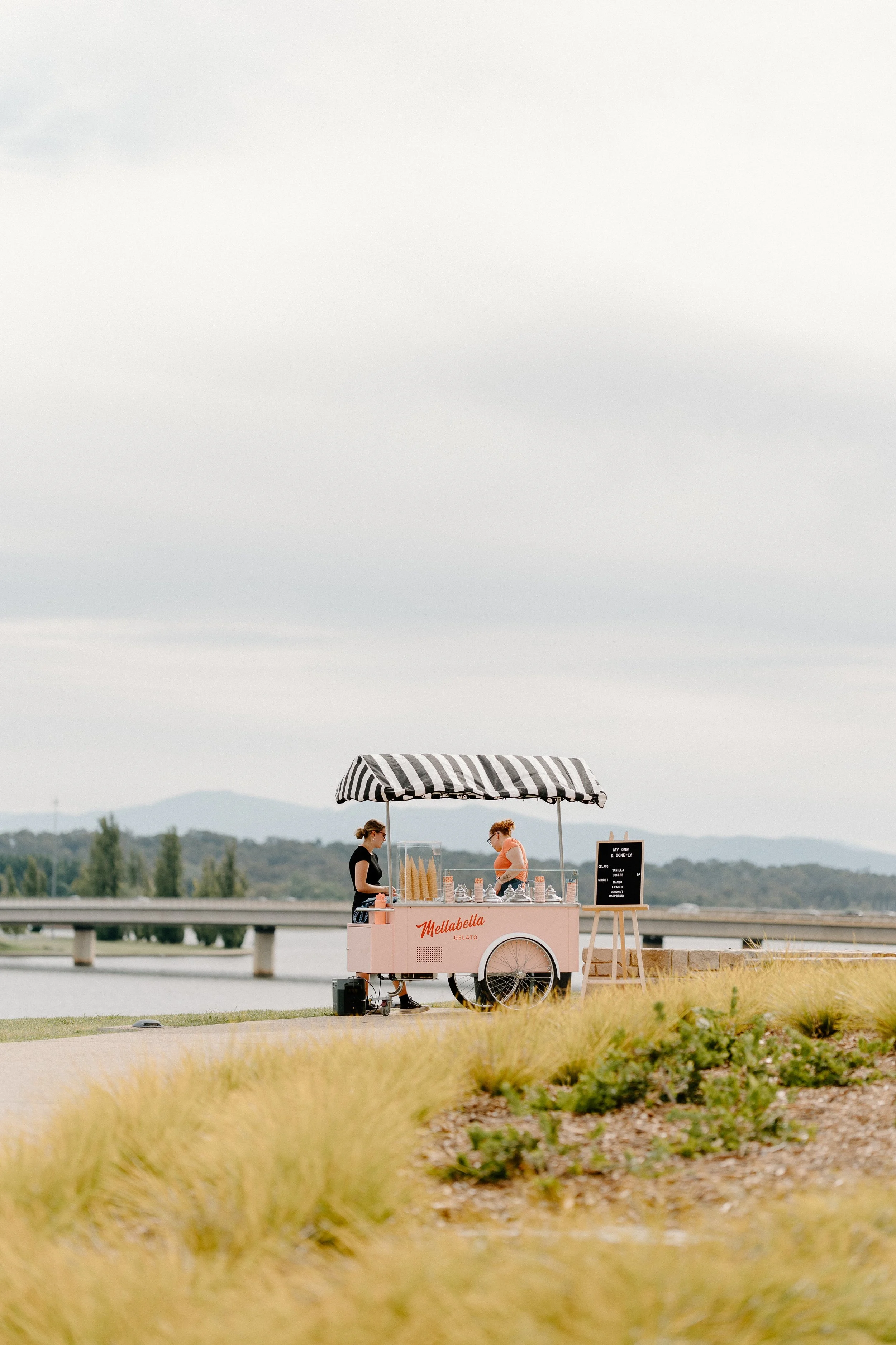 A pink ice cream cart with a black and white striped canopy by a river, with two women attending it and a menu board displayed nearby, against a backdrop of mountains and a cloudy sky.