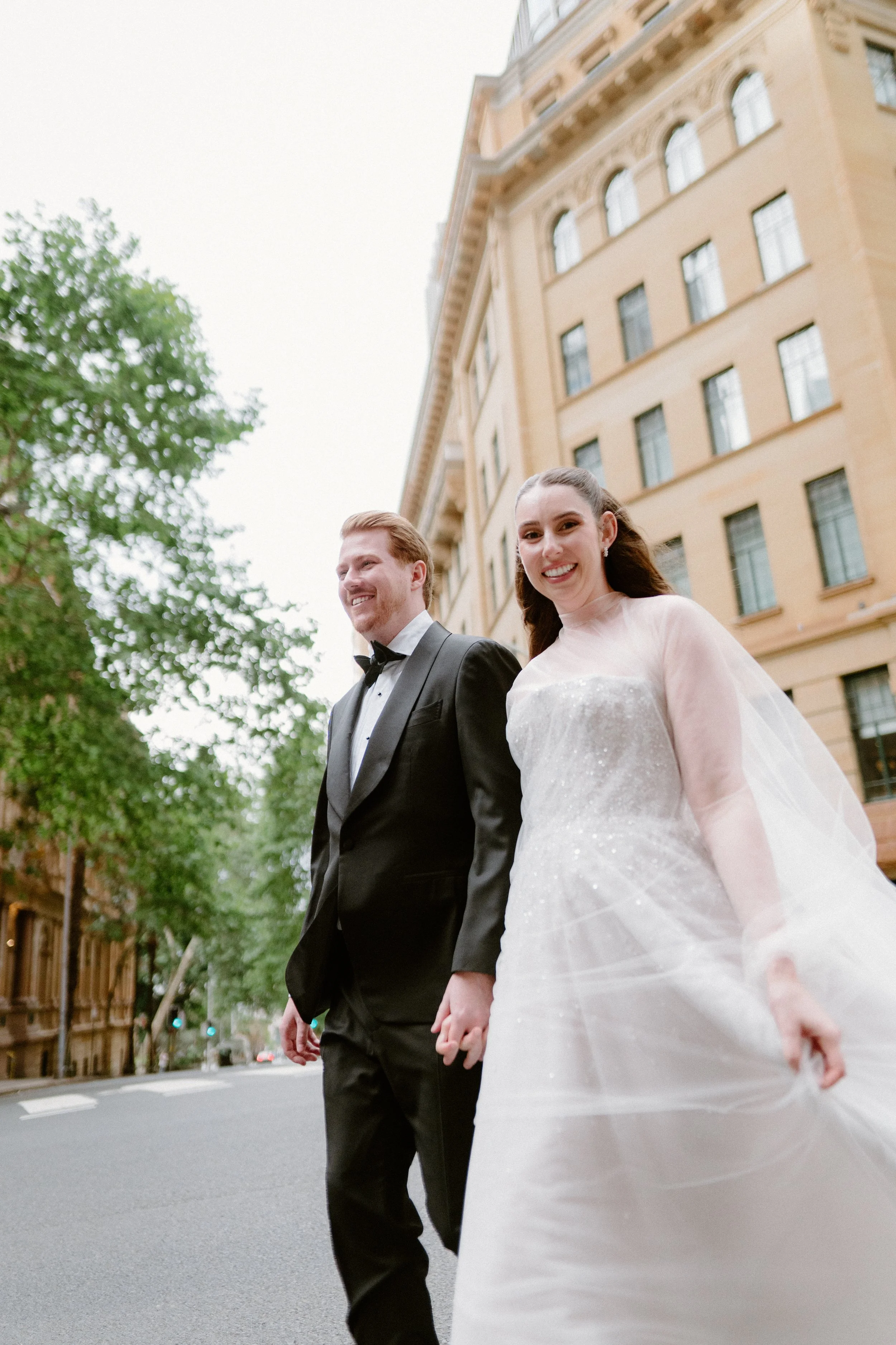 A bride and groom walking hand in hand outdoors in front of a large beige building, smiling, on a city street with trees.