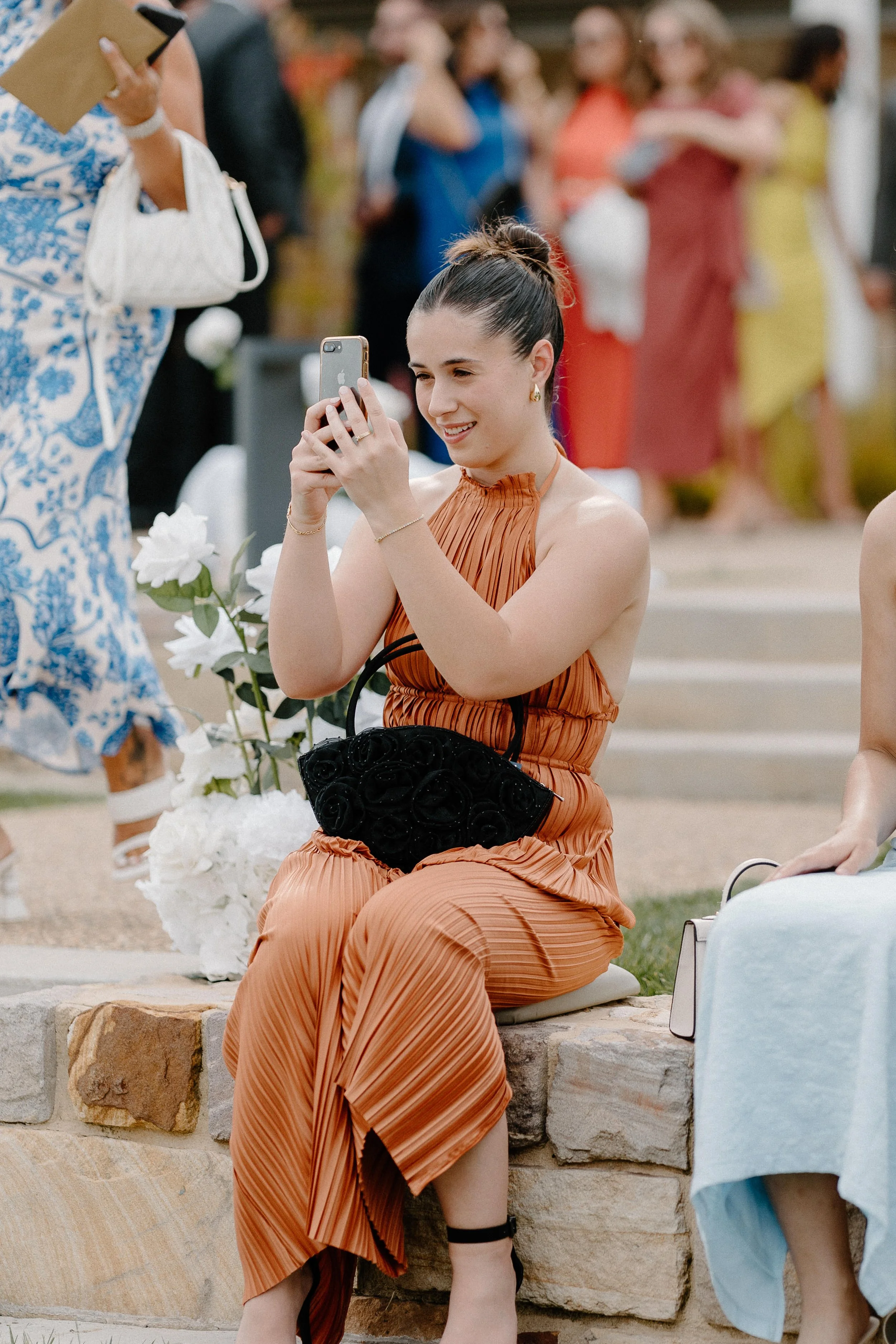 A young woman in a rust-colored pleated dress is sitting on a stone ledge at an outdoor event, taking a photo with her smartphone, with people mingling in the background.