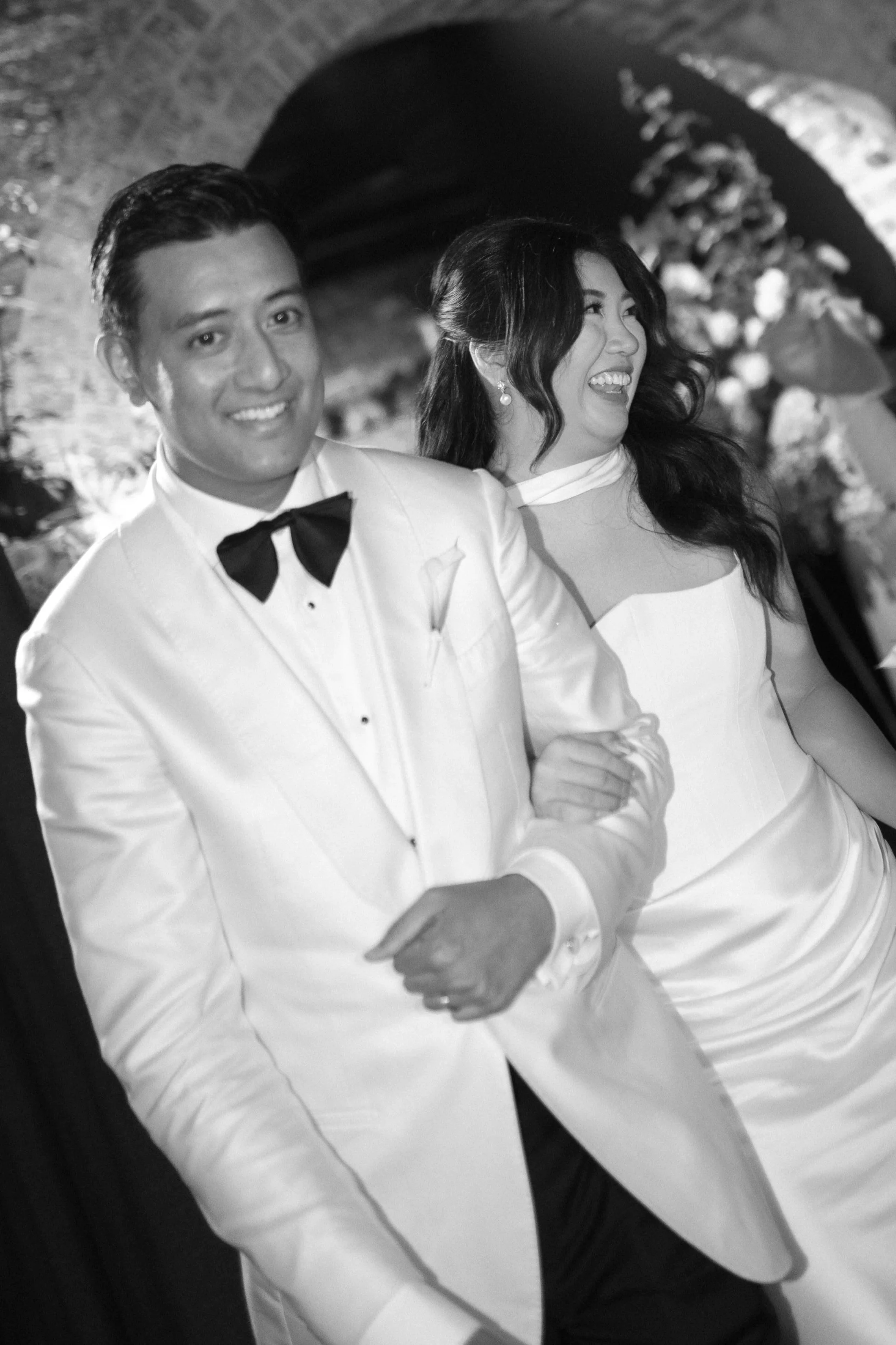 A black-and-white photo of a happy couple at a wedding, walking arm-in-arm in an indoor venue with a brick archway in the background. The man is wearing a white tuxedo jacket with a black bow tie, and the woman is in a white dress with a sheer neckli