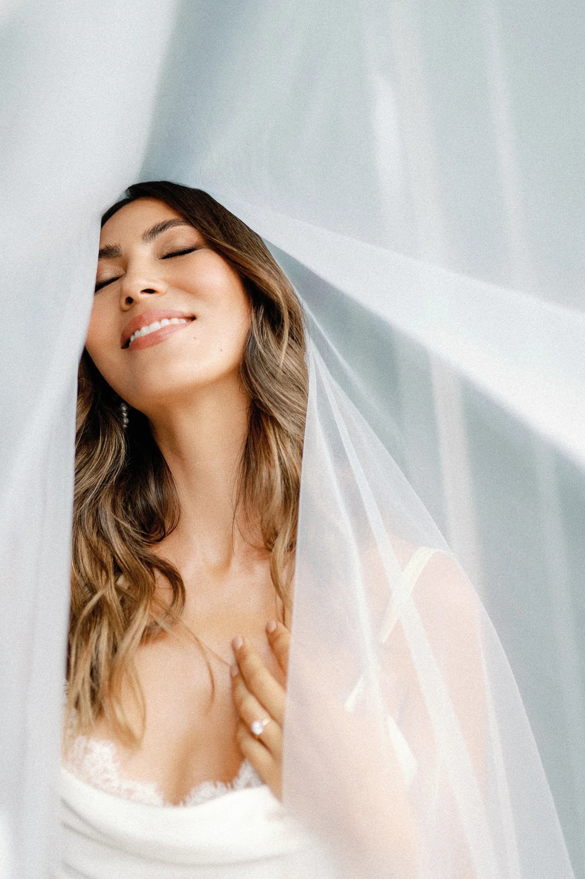 A woman with long, wavy hair smiling with her eyes closed, wearing a white lingerie and a veil, in a softly lit room.