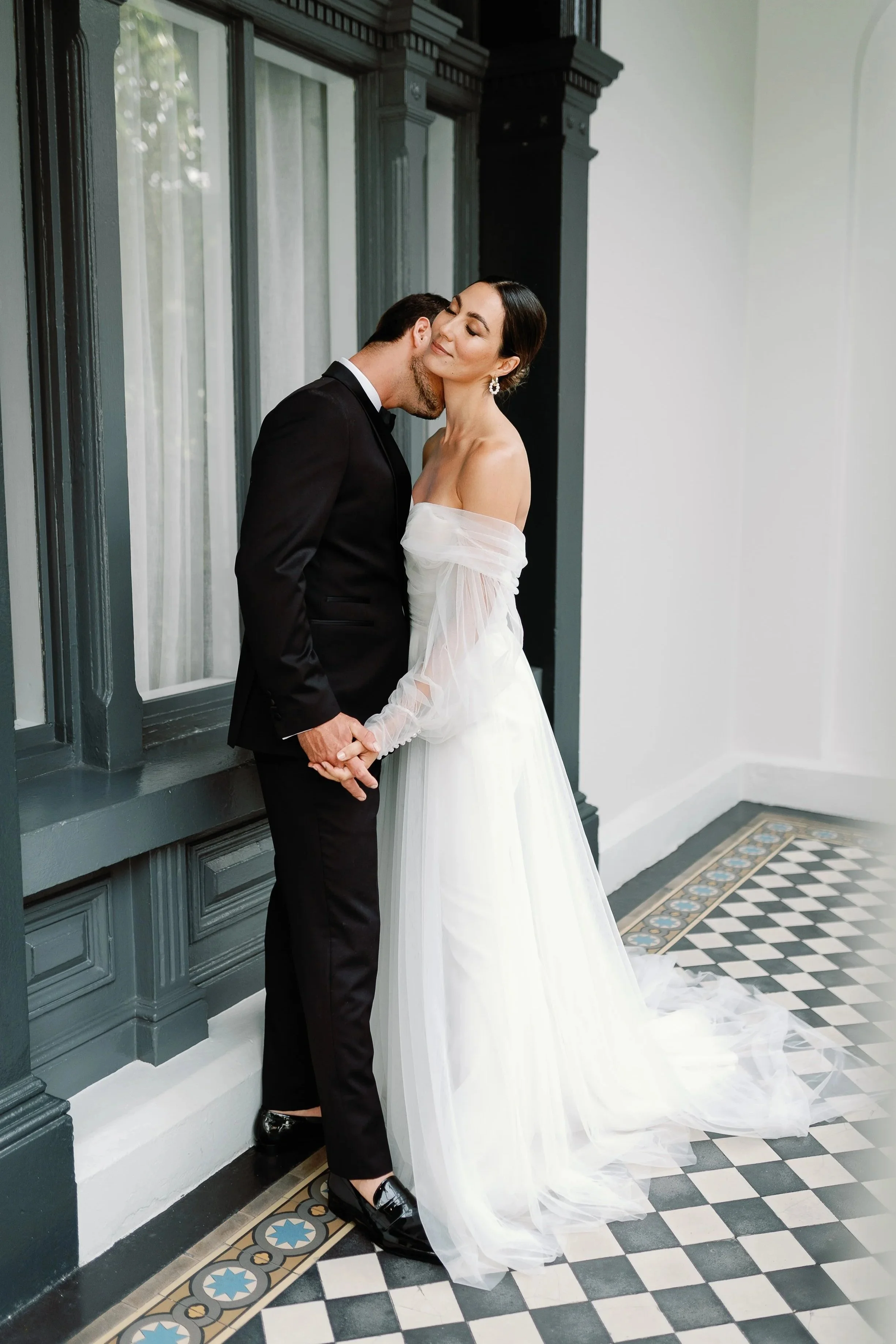 A bride and groom holding hands, sharing an intimate moment in a historic building with decorative black and white tile flooring.