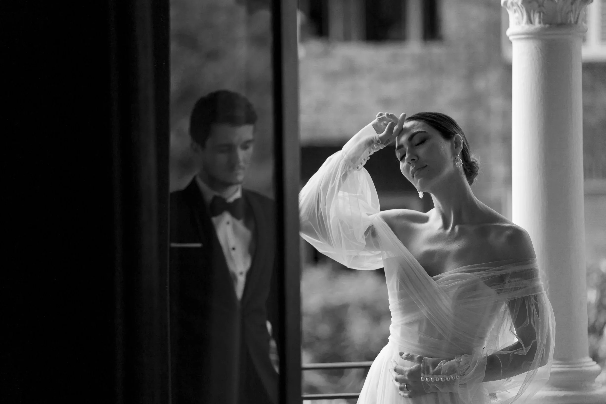 A black and white photo of a woman in an off-shoulder dress with sheer sleeves standing outdoors near a column, with her eyes closed and hand on her forehead, while a man in a tuxedo with a bow tie looks downwards from behind a glass or mirror.
