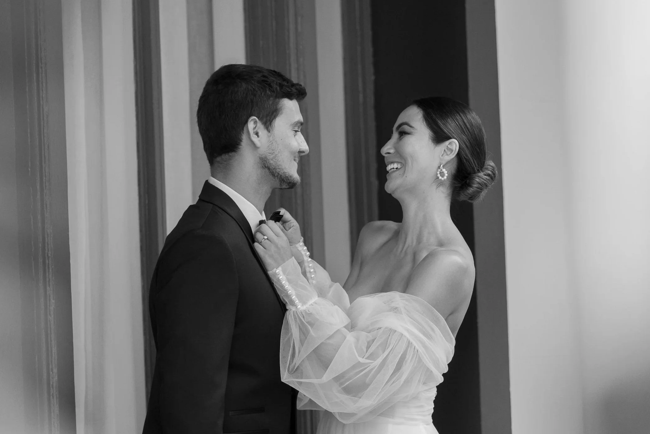 A black and white photo of a bride and groom smiling at each other during their wedding, with the bride adjusting the groom's bow tie.