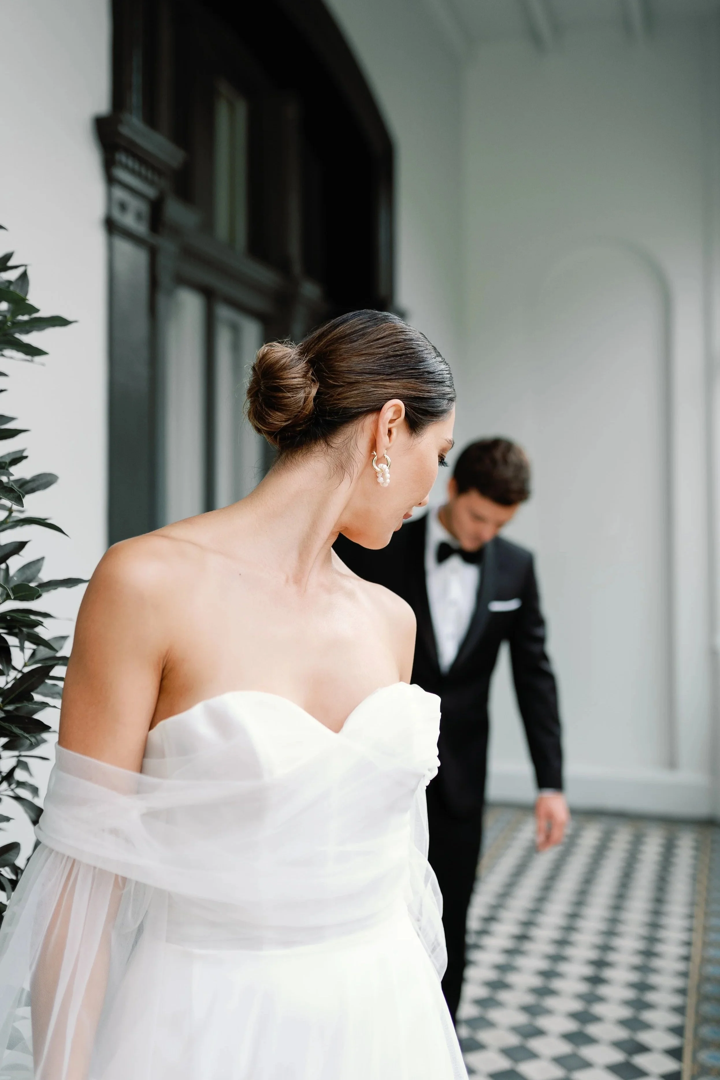 Bride with elegant updo and pearl earrings, wearing a strapless white wedding gown with sheer detailing, looking down, with groom in tuxedo adjusting something in the background, on a patterned black and white floor in a bright room.