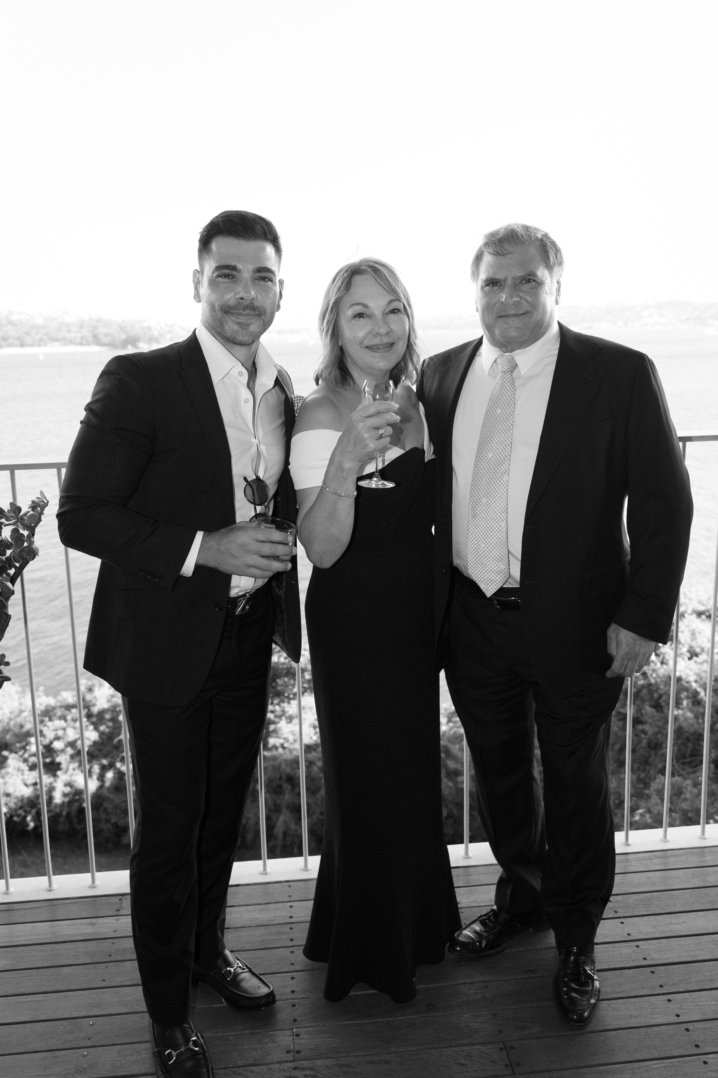 Black and white photo of three people in formal attire holding drinks on a balcony with water and hills in the background.
