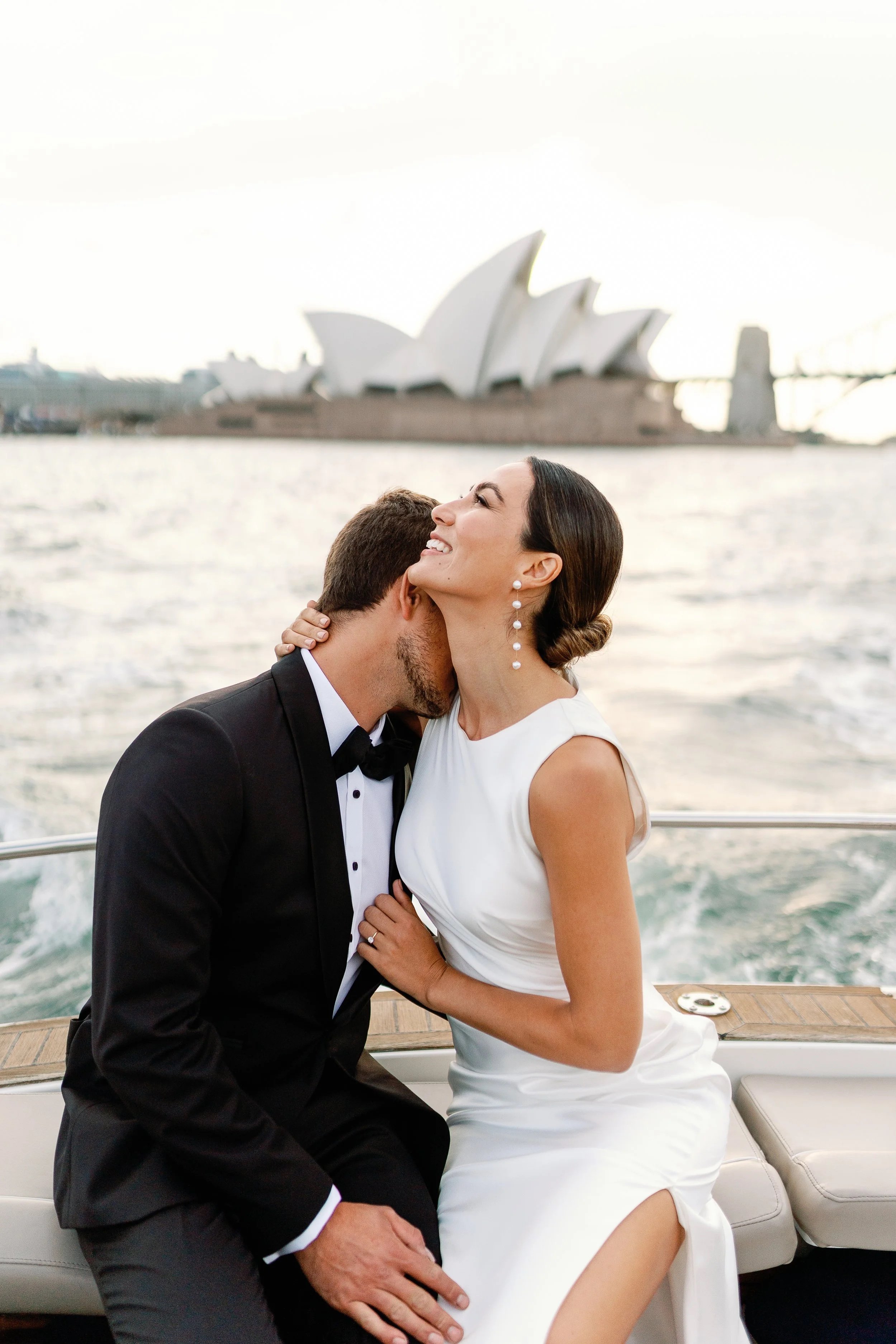 A couple in a wedding dress and tuxedo sharing an intimate moment on a boat, with the Sydney Opera House in the background.
