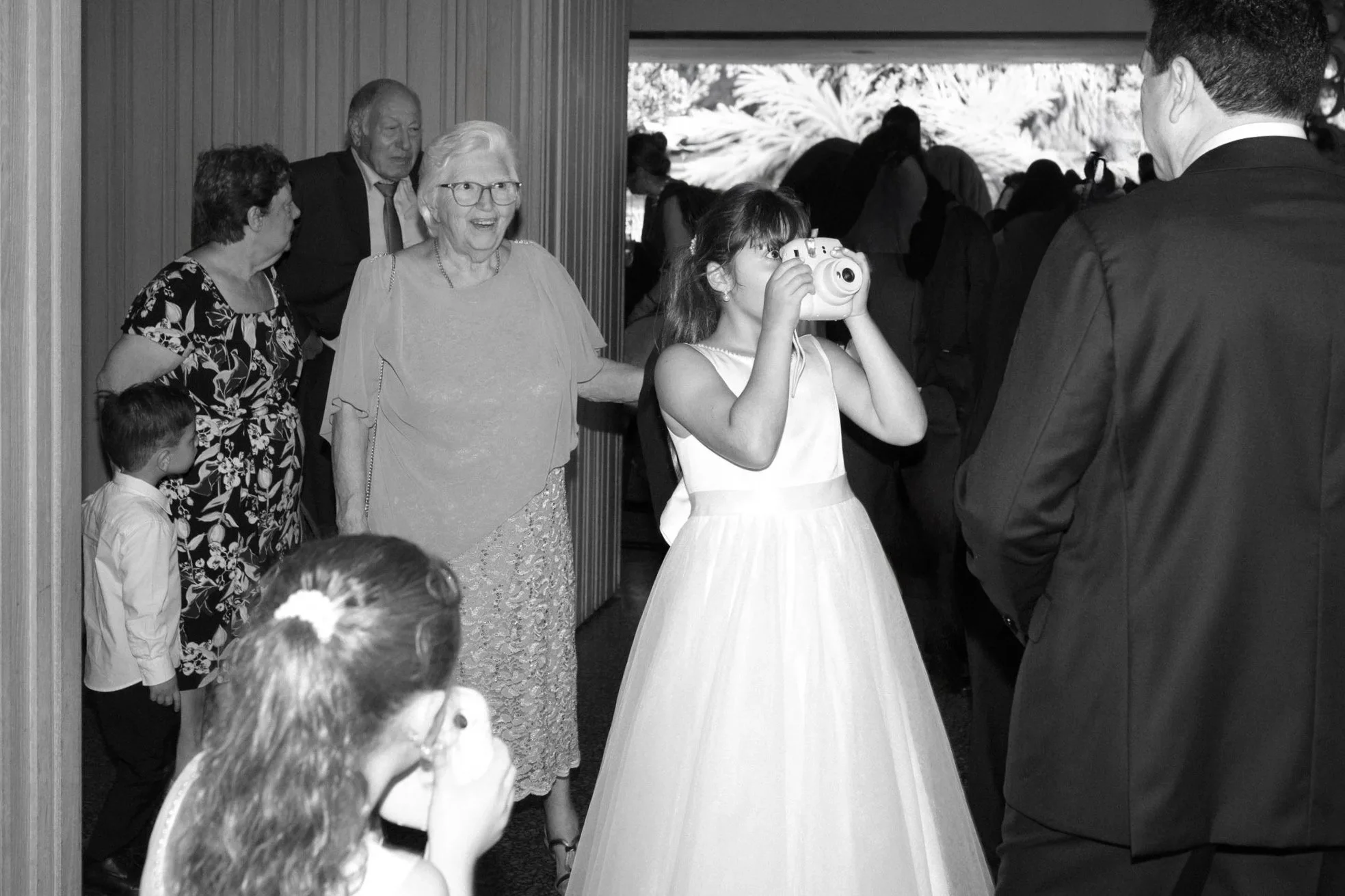 A young girl in a white dress taking a photo with a camera at a social event, surrounded by adults and children. Some people are smiling and observing the girl, while others are gathered in a room with a view of outdoors with palm trees.