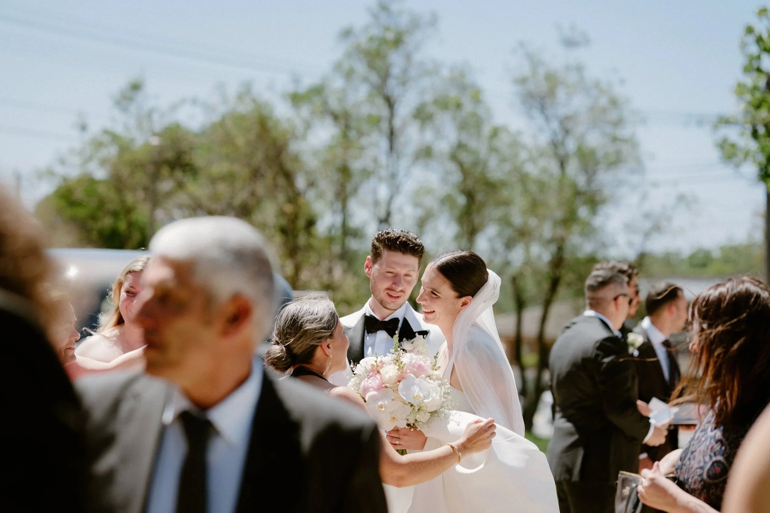 A bride and groom at their wedding, smiling and holding a bouquet, outdoors with trees in the background, surrounded by guests in formal attire.