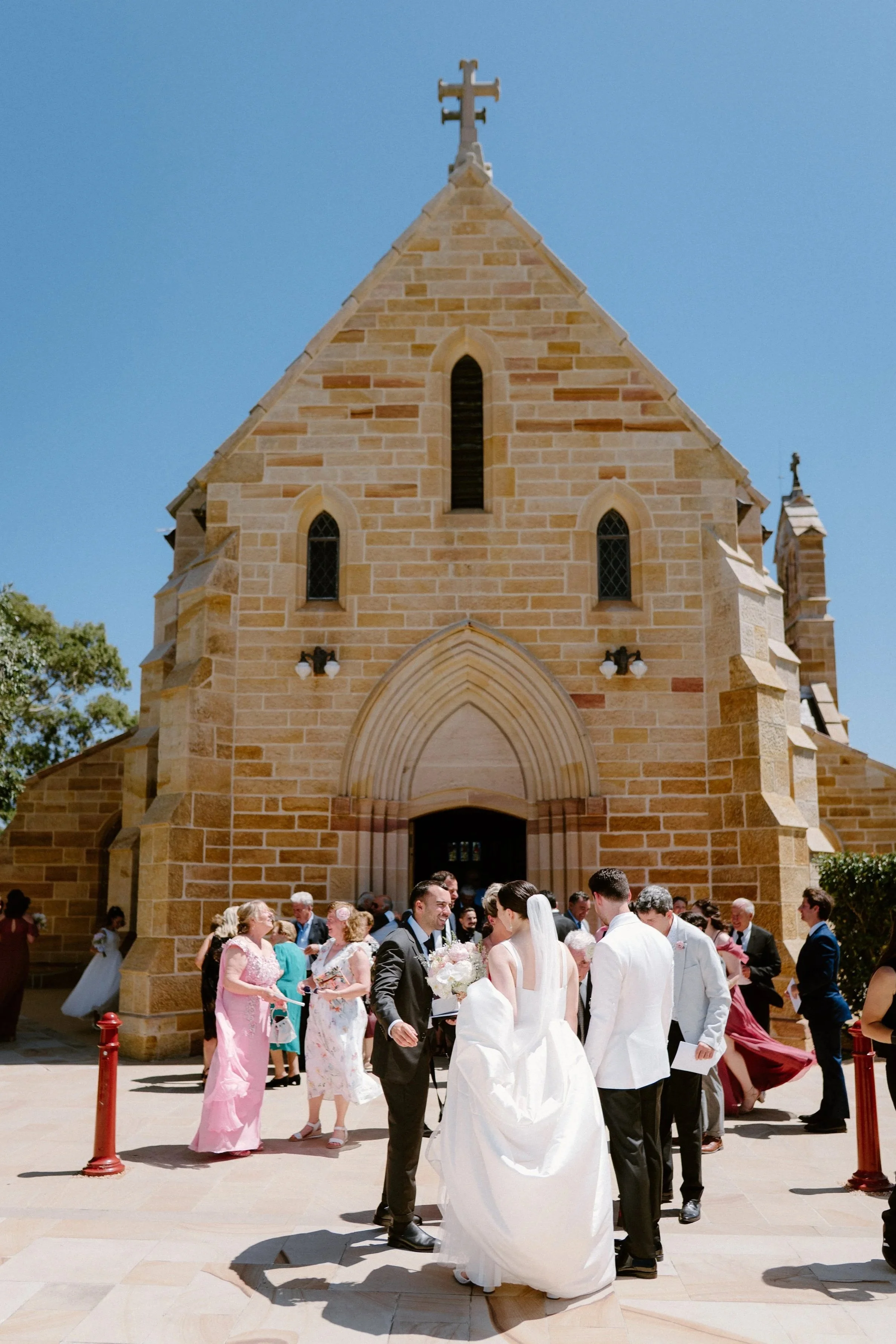 A wedding outside a church with a brick facade and a cross on top, with the bride and groom surrounded by guests on a sunny day.