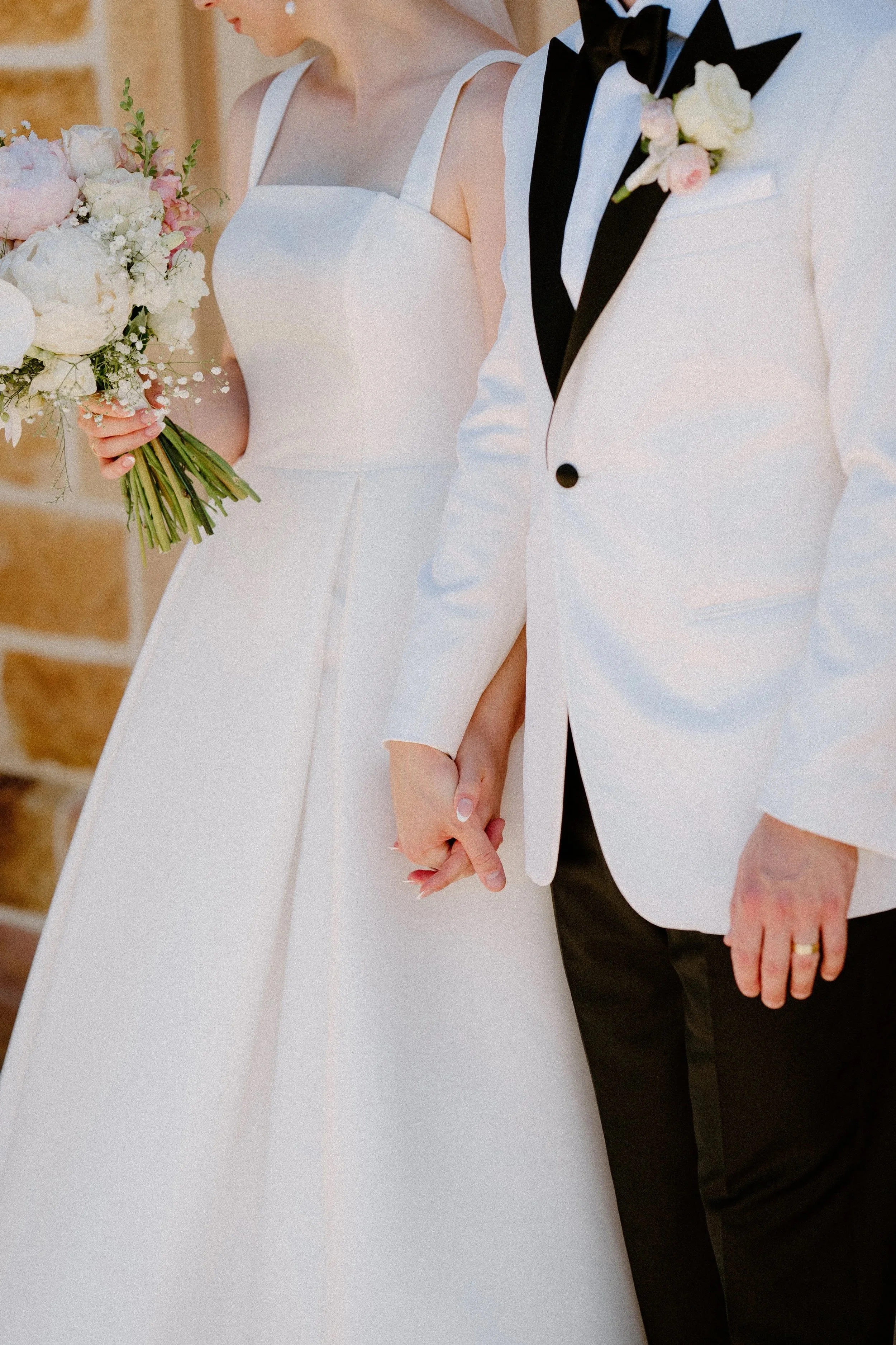 Close-up of a bride and groom holding hands during their wedding, with the bride wearing a white dress and holding a bouquet, and the groom dressed in a white tuxedo jacket with a black bow tie and boutonniere.