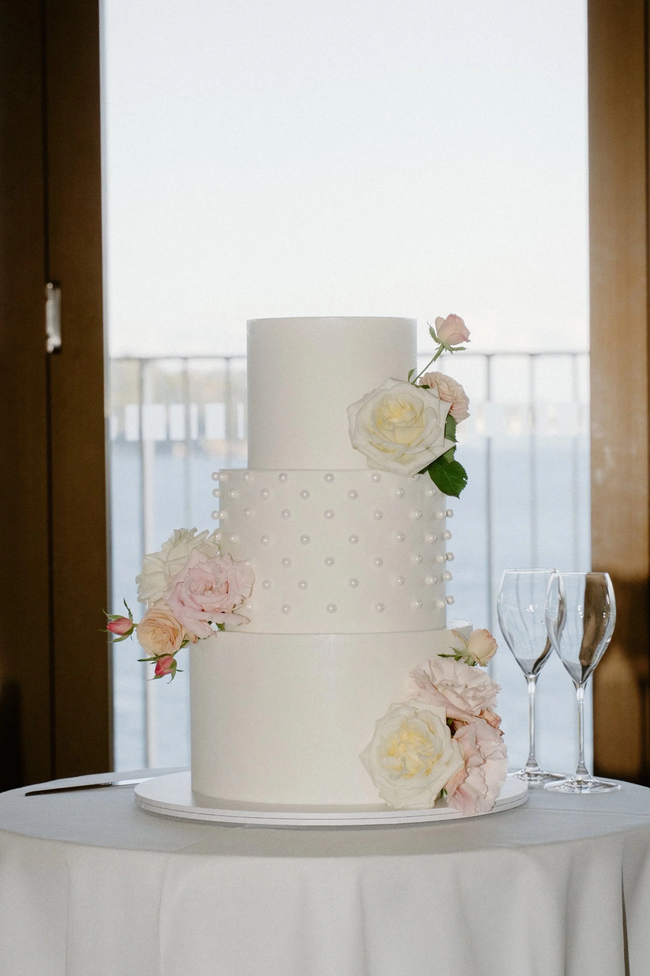 Three-tiered white wedding cake decorated with white and pink roses and small white pearls, placed on a white tablecloth-covered table with two empty champagne flutes to the right.