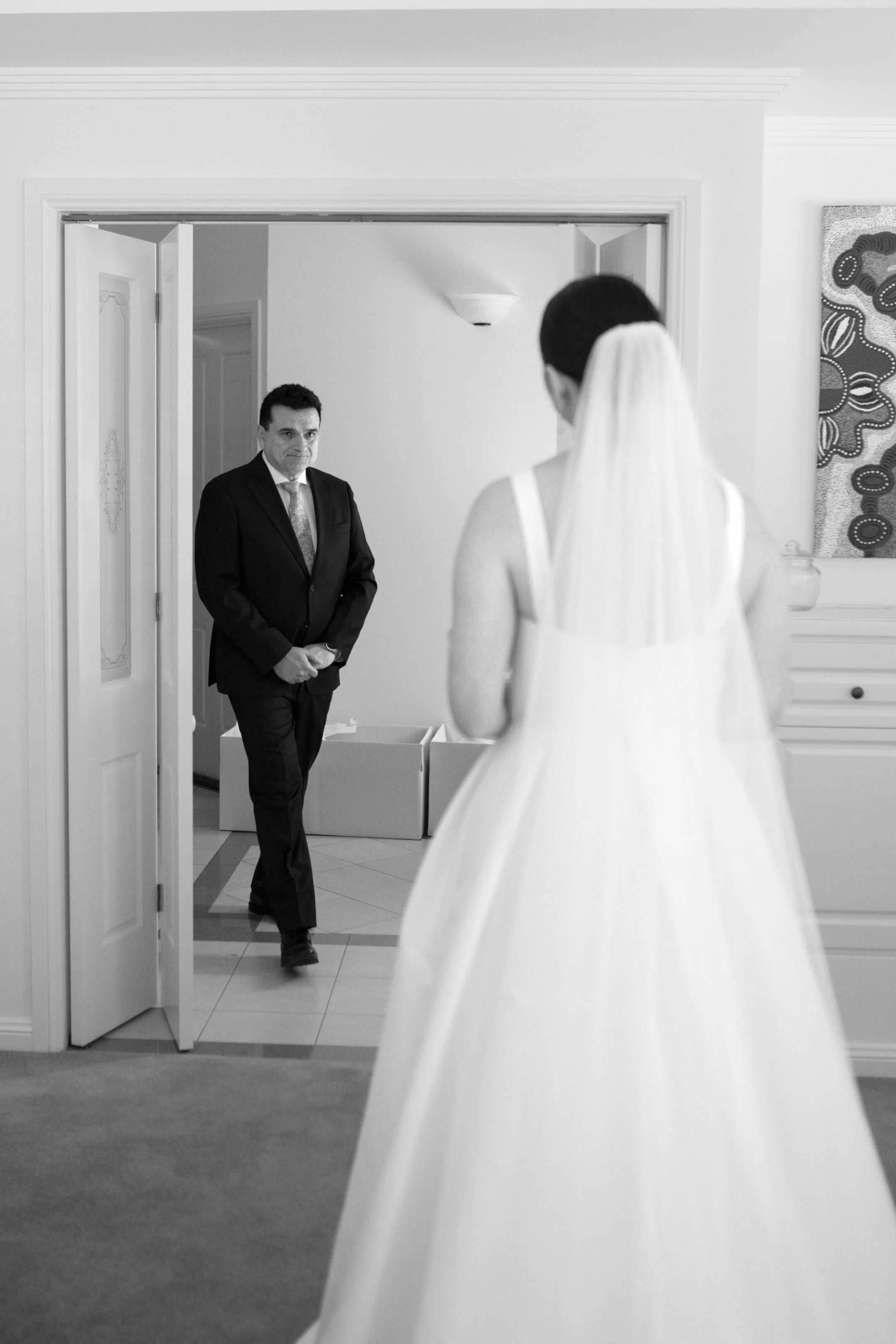 A bride in a white wedding dress with a veil is facing a man in a suit, standing in a doorway inside a room.