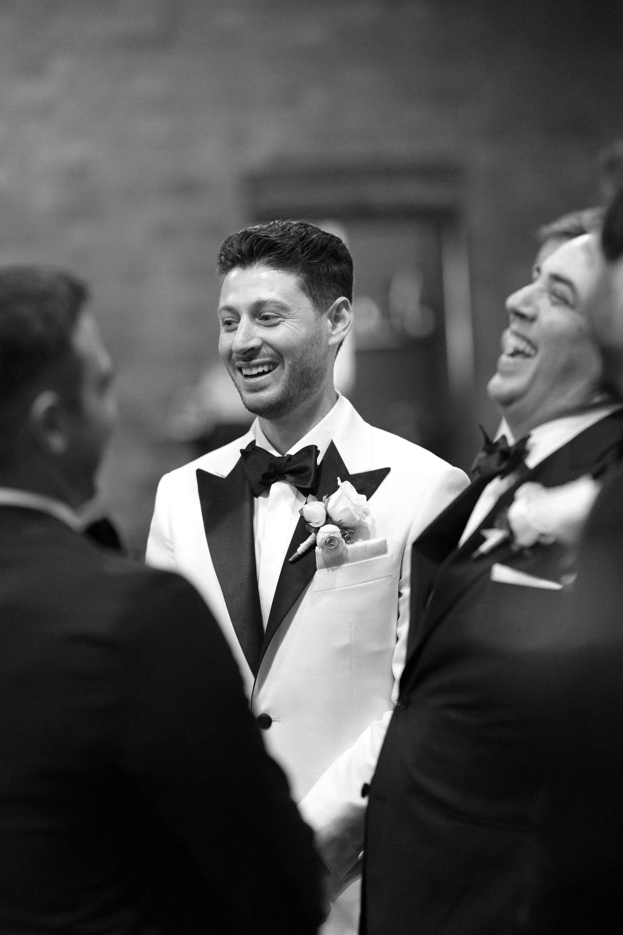 Three men in tuxedos smiling and laughing during a wedding ceremony, with the groom in the center.