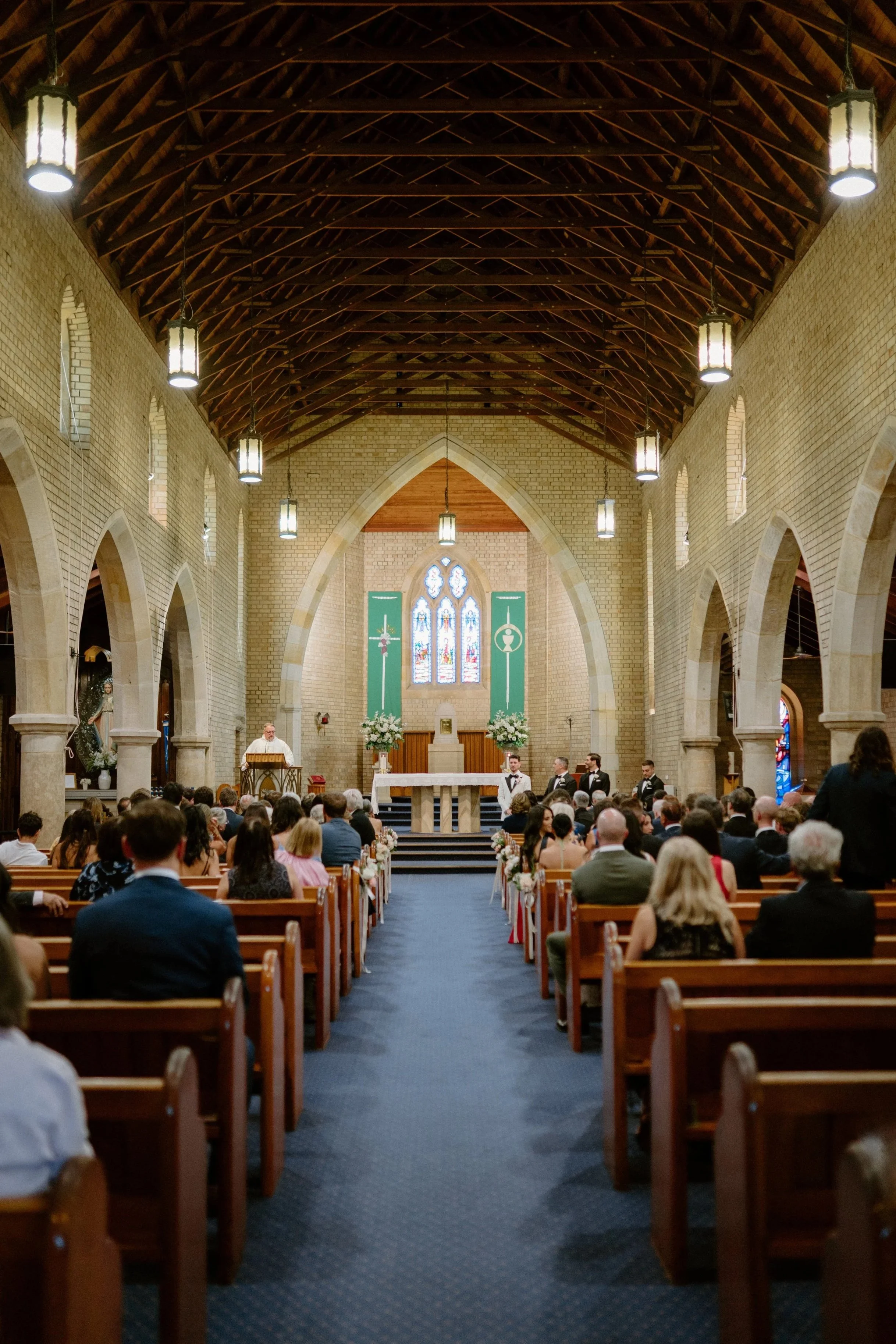 Interior of a church decorated for a wedding ceremony with guests seated, a priest officiating, and brides and groom standing at the altar.