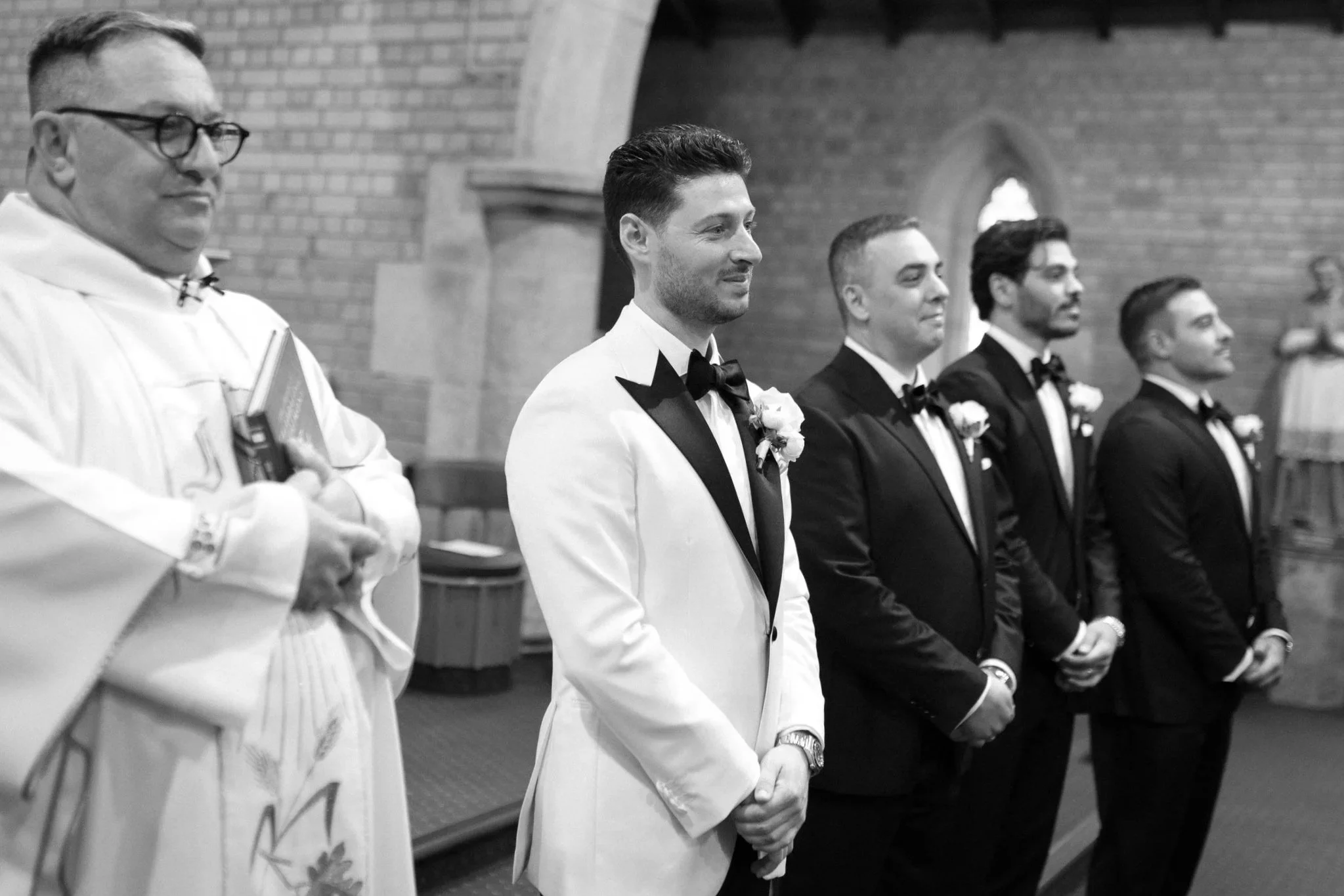 Groom and groomsmen standing in a church during a wedding ceremony, wearing tuxedos with boutonnières, with a priest in the foreground holding a book.