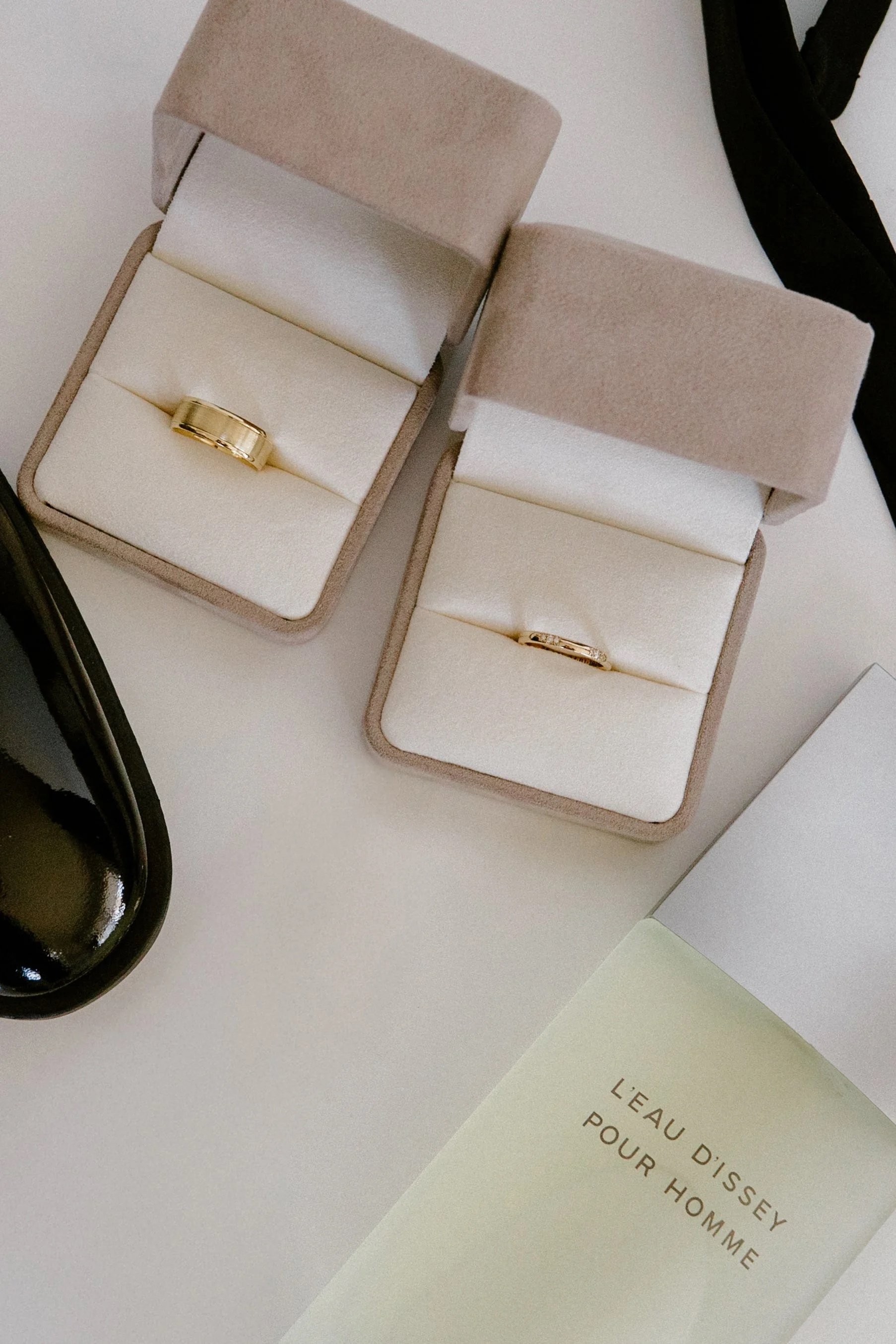 Two beige jewelry boxes with white inserts, each containing a gold ring, placed on a white surface. There is a black object near the boxes and a partially visible white box with text that reads "L'EAU D'ISSEY POUR HOMME."