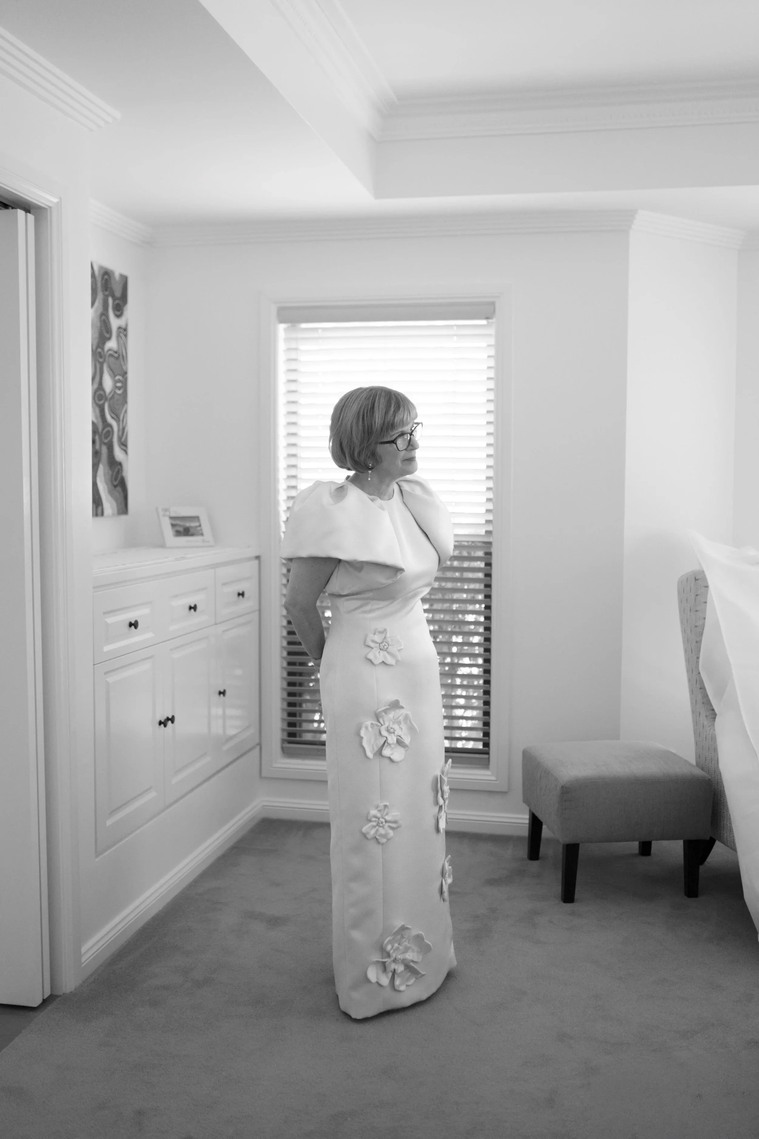 An older woman with short hair and glasses wearing a long white dress with floral appliqué, standing in a bright room with blinds, white cabinets, and a chair.