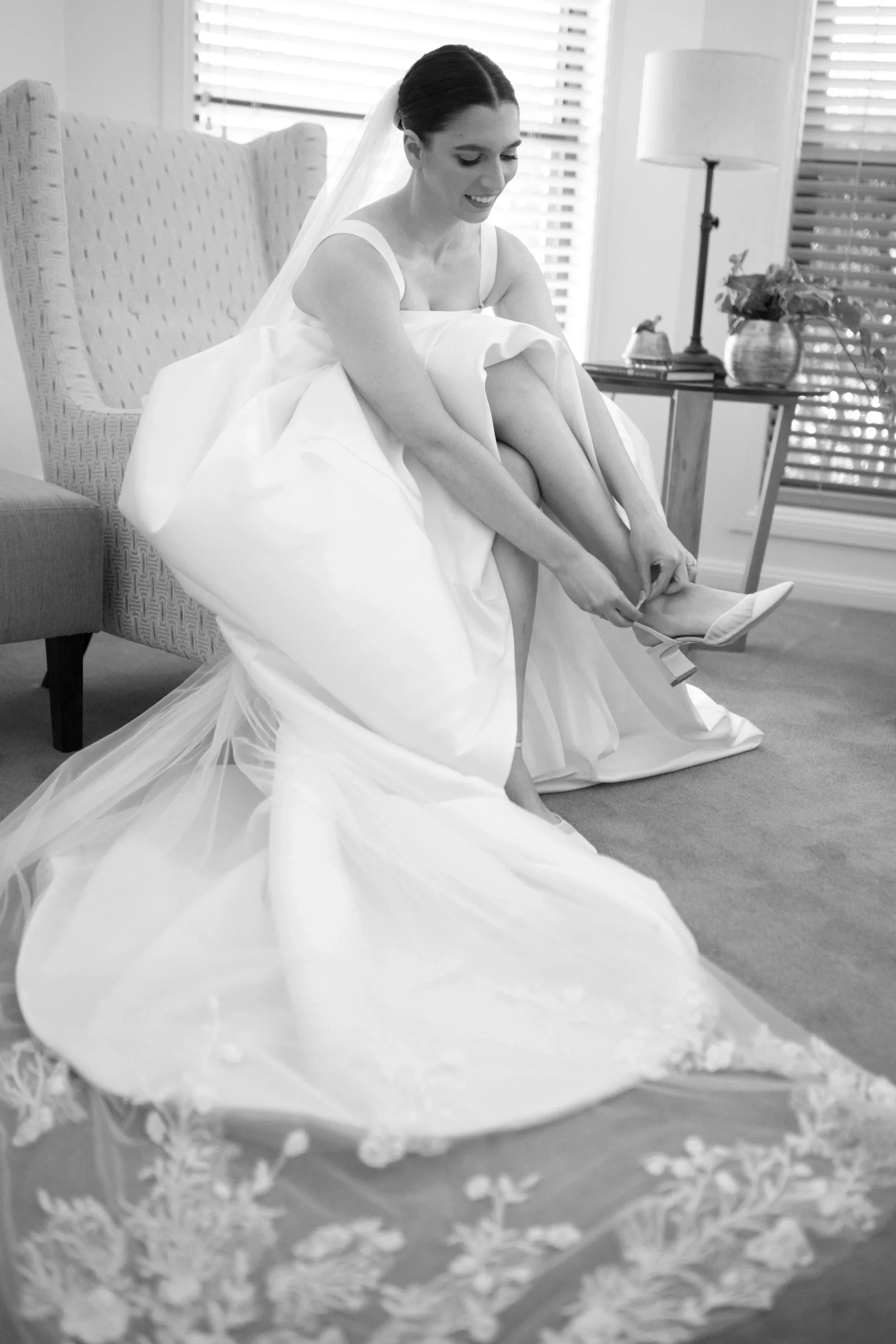 A woman in a wedding dress sitting on a chair, putting on high heel shoes, in a room with wooden blinds and a side table with a lamp and a potted plant.