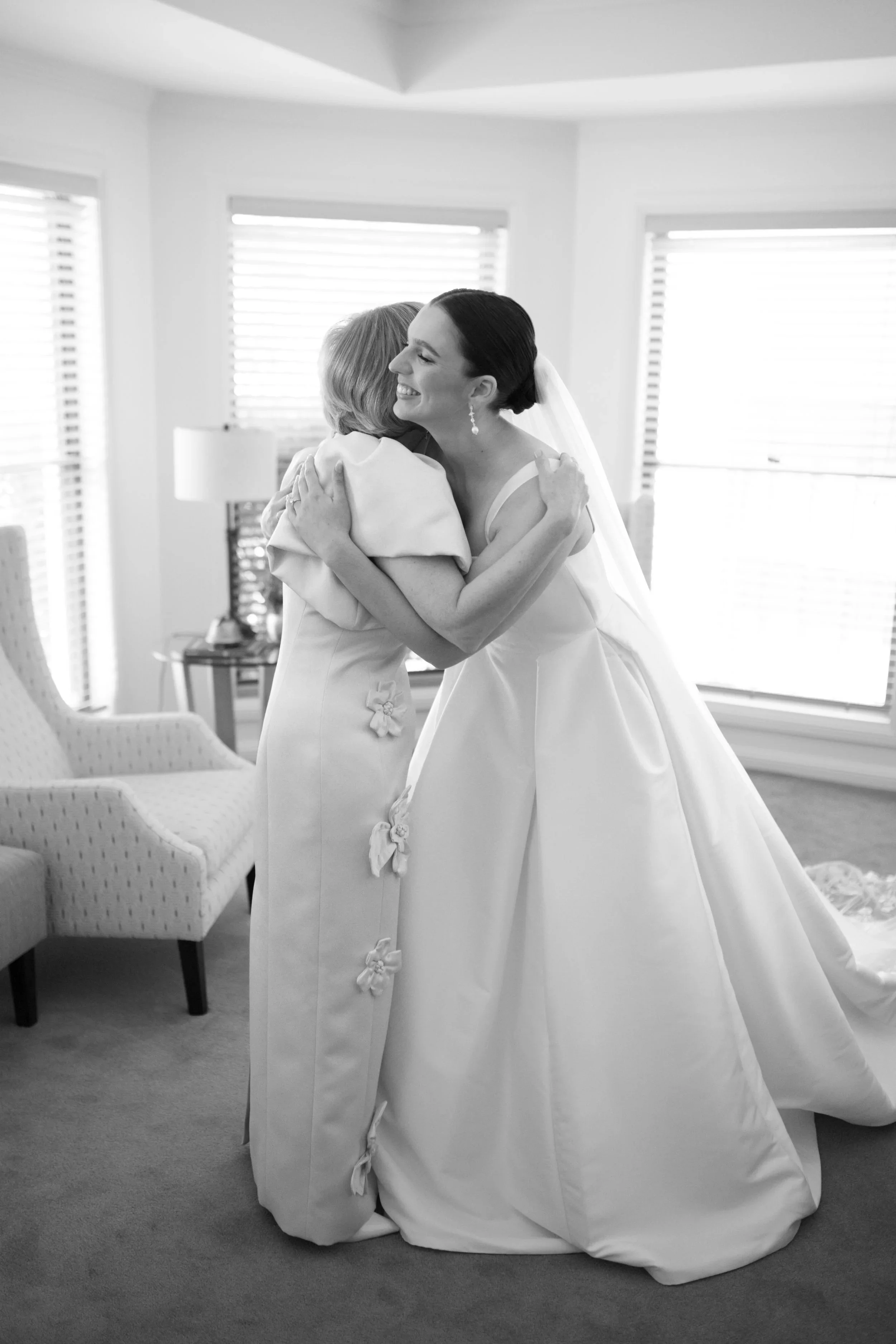 A bride in a white wedding gown hugging an older woman in a bright room with large windows.