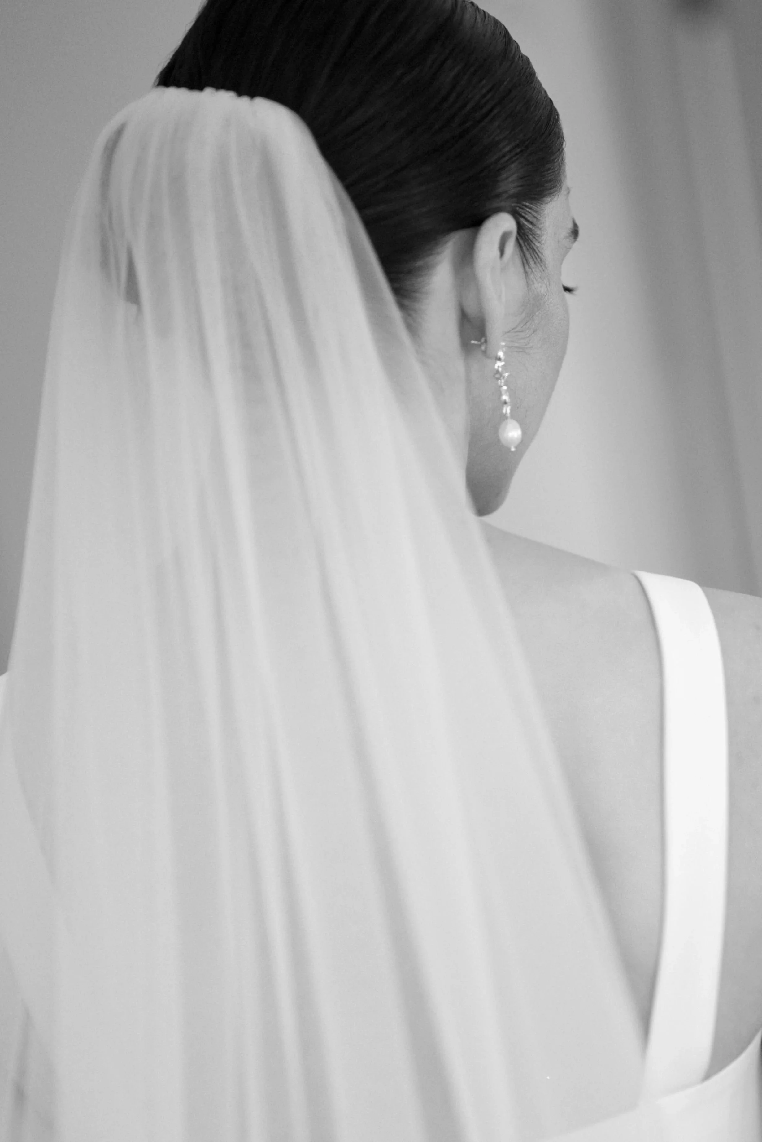 Back view of a bride with a wedding veil and pearl earrings, wearing a white dress.