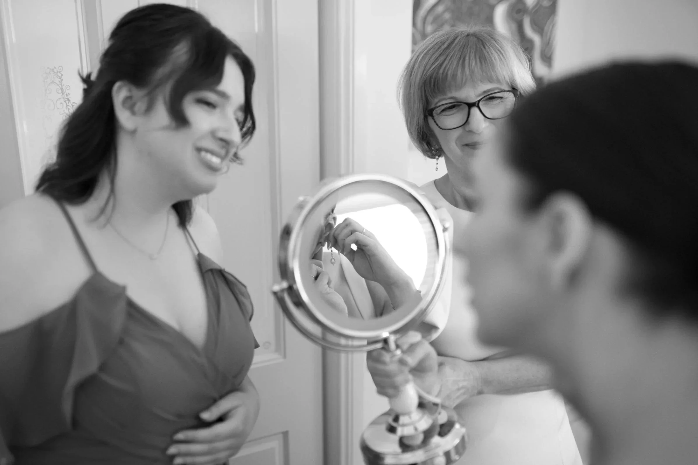 Three women standing in front of a mirror; one woman is holding a small framed mirror, and all are smiling and looking at each other, in a domestic setting.