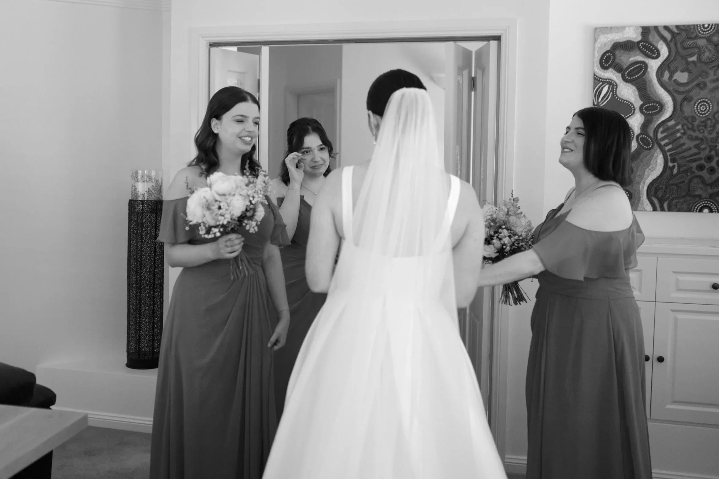 A bride in a white wedding dress and veil is seen from the back, facing four women who are likely her bridesmaids, dressed in matching dresses, with two holding bouquets, inside a room with modern decor and an abstract painting on the wall.
