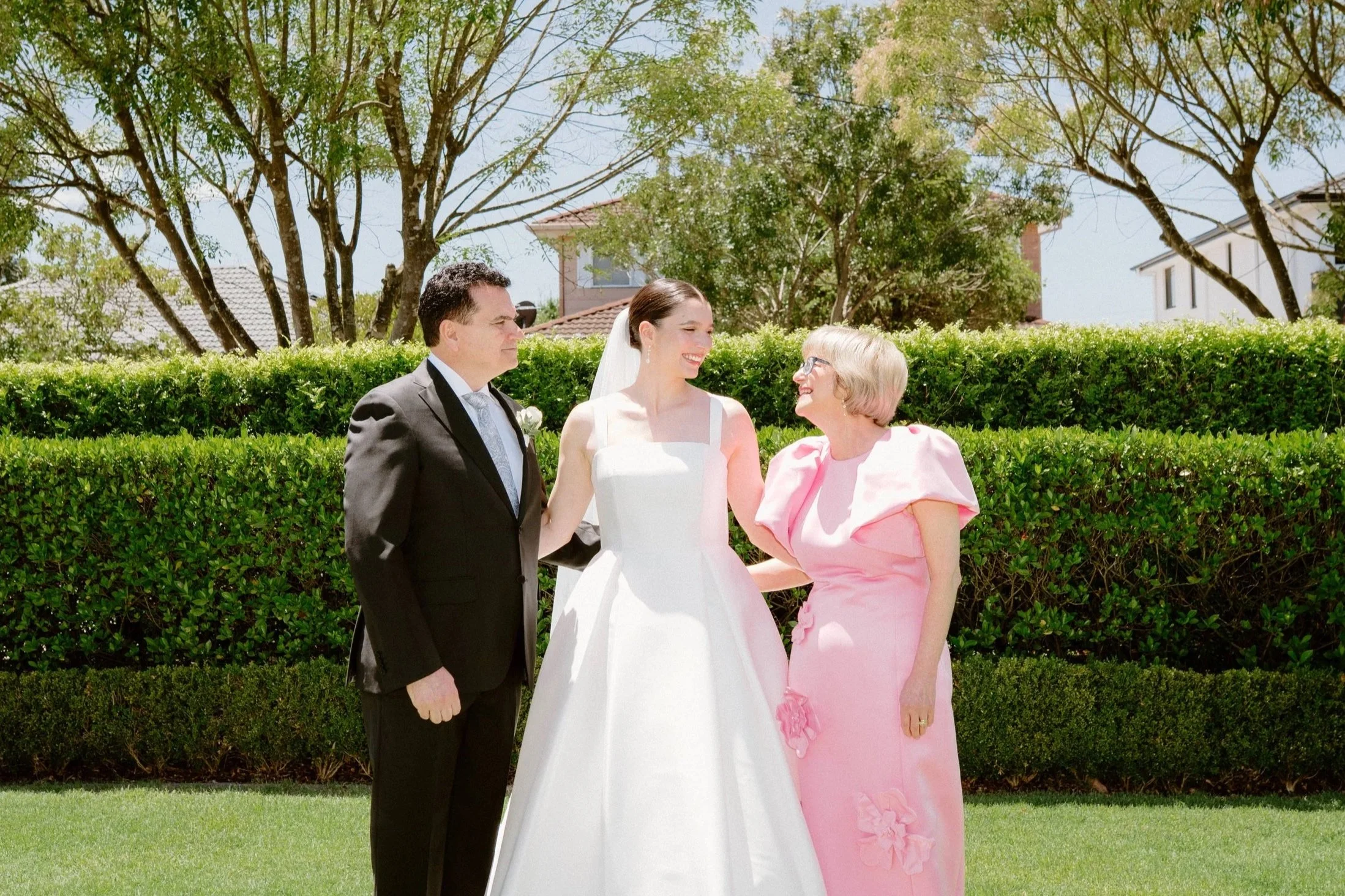 A wedding scene outdoors with a bride, groom, and an older woman in pink dress smiling and talking