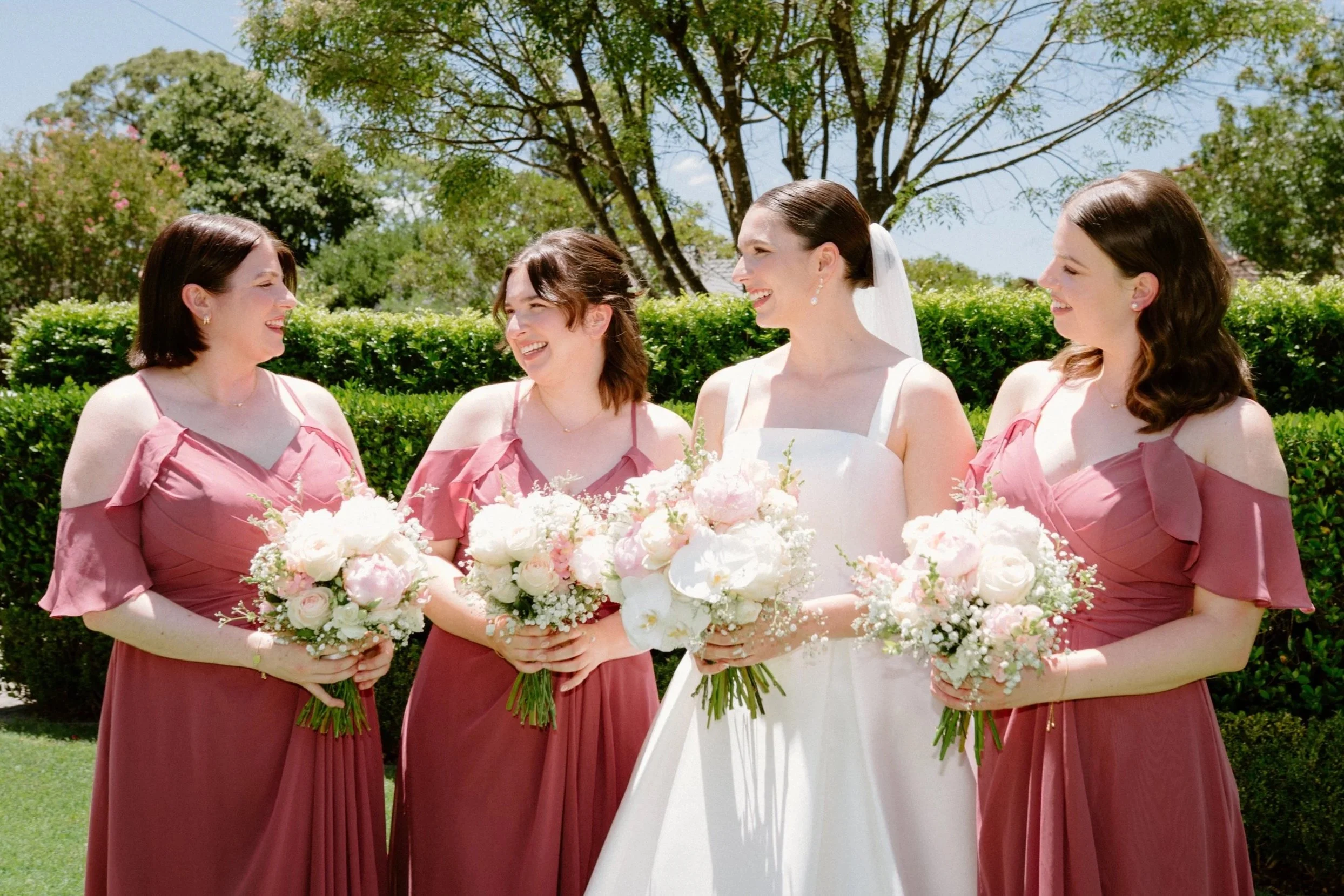 A bride in a white wedding dress and four bridesmaids in mauve dresses holding bouquets, standing outdoors on a sunny day with trees and greenery in the background.