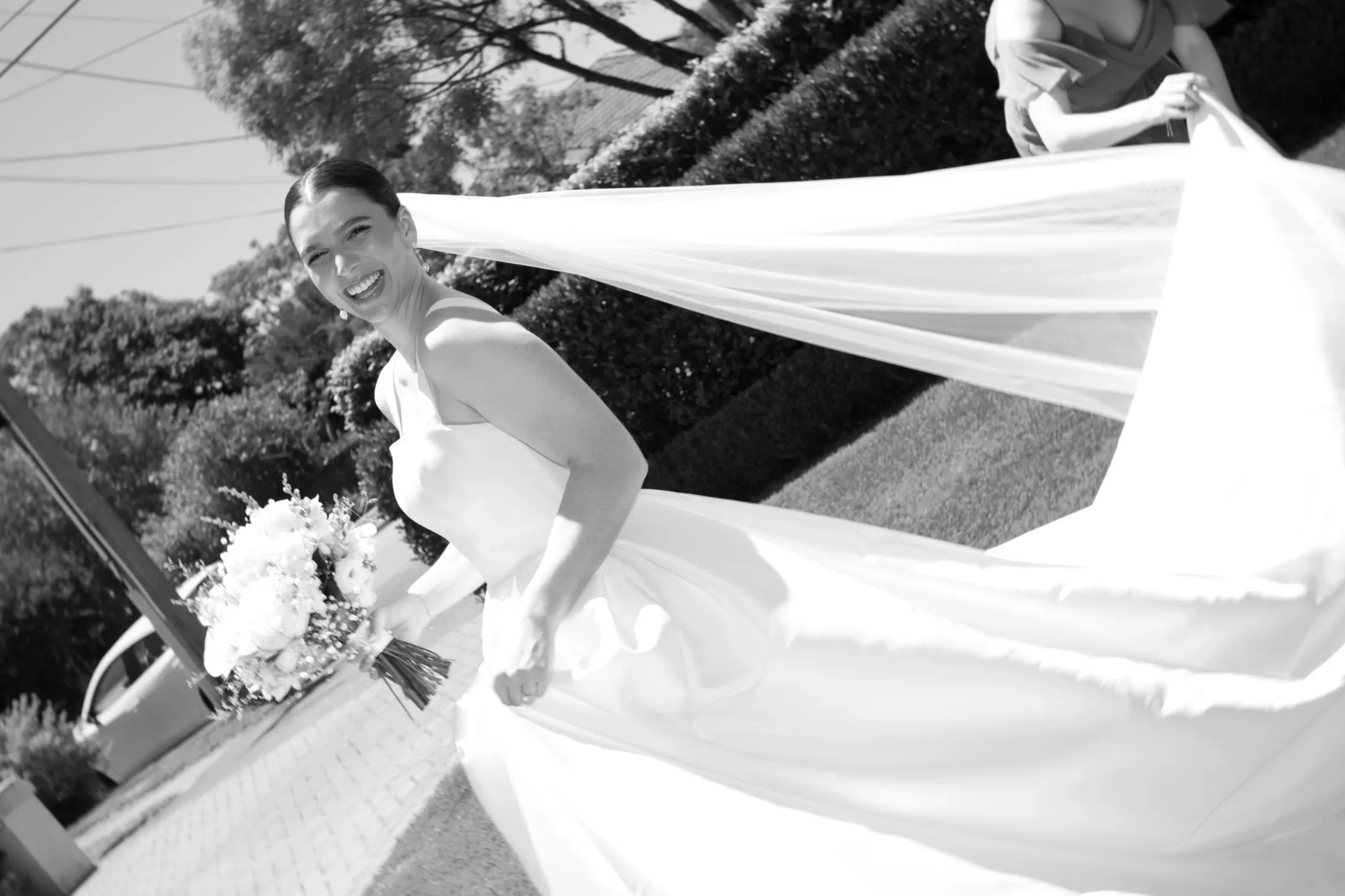 A woman in a wedding dress holding a bouquet of flowers, smiling outdoors during daytime.