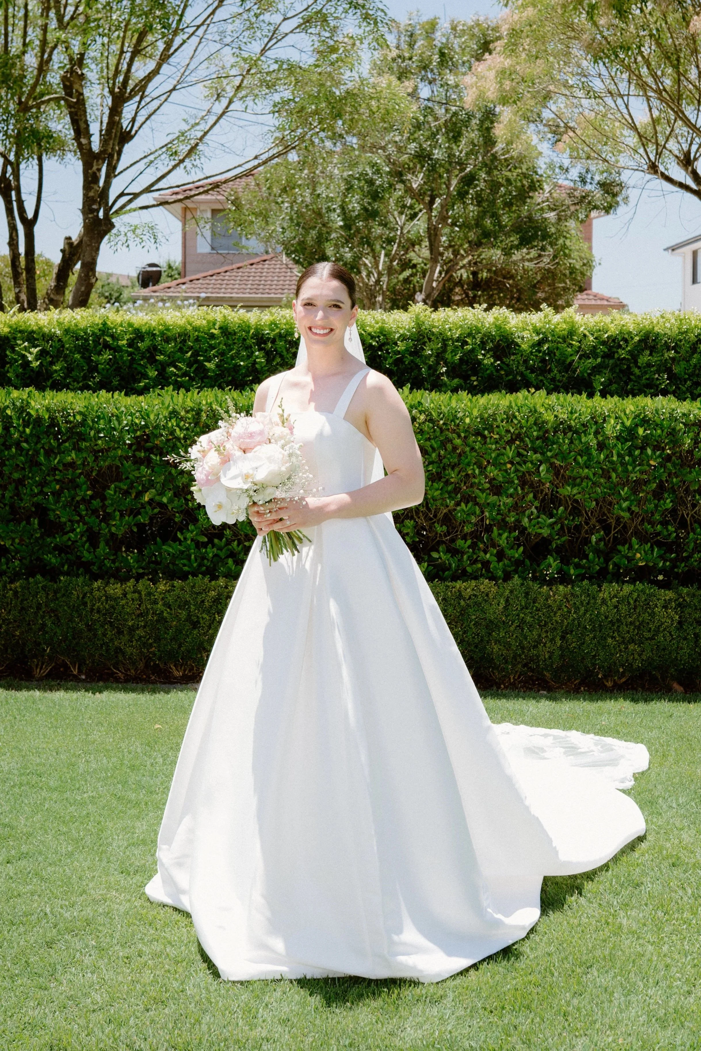 Bride in a white wedding dress holding a bouquet of pink and white flowers outdoors on a bright, sunny day.