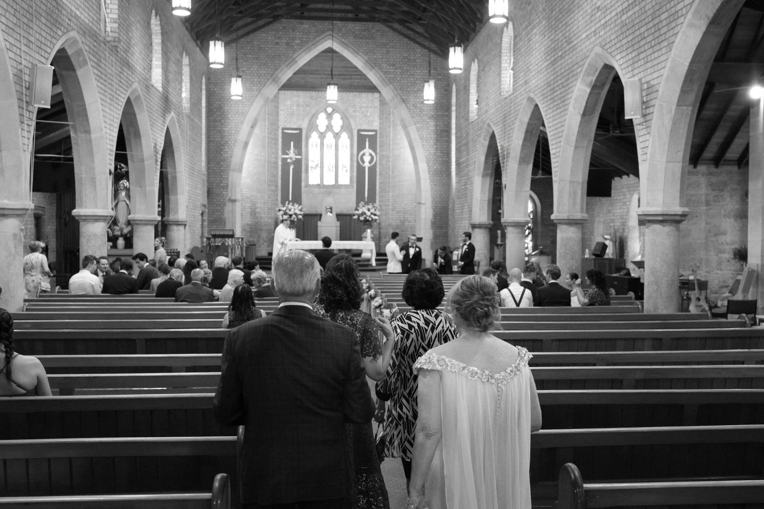 Black and white photo of a wedding ceremony inside a church with arched ceilings. The couple is at the altar, with guests seated in pews watching. The scene depicts a religious wedding with a priest officiating.