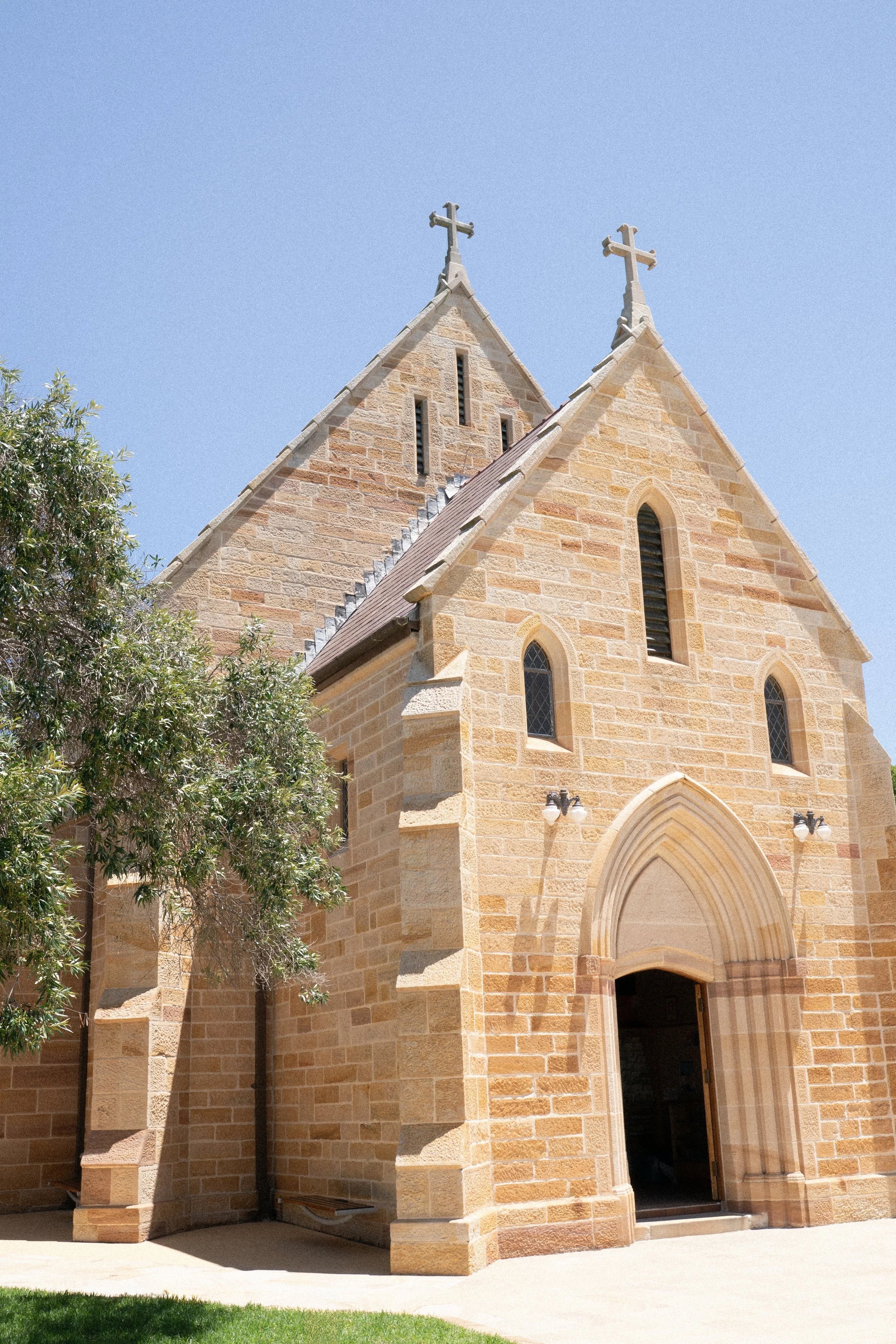 A stone church with two steeples topped with crosses, surrounded by a small grassy area and a tree, under a clear blue sky.