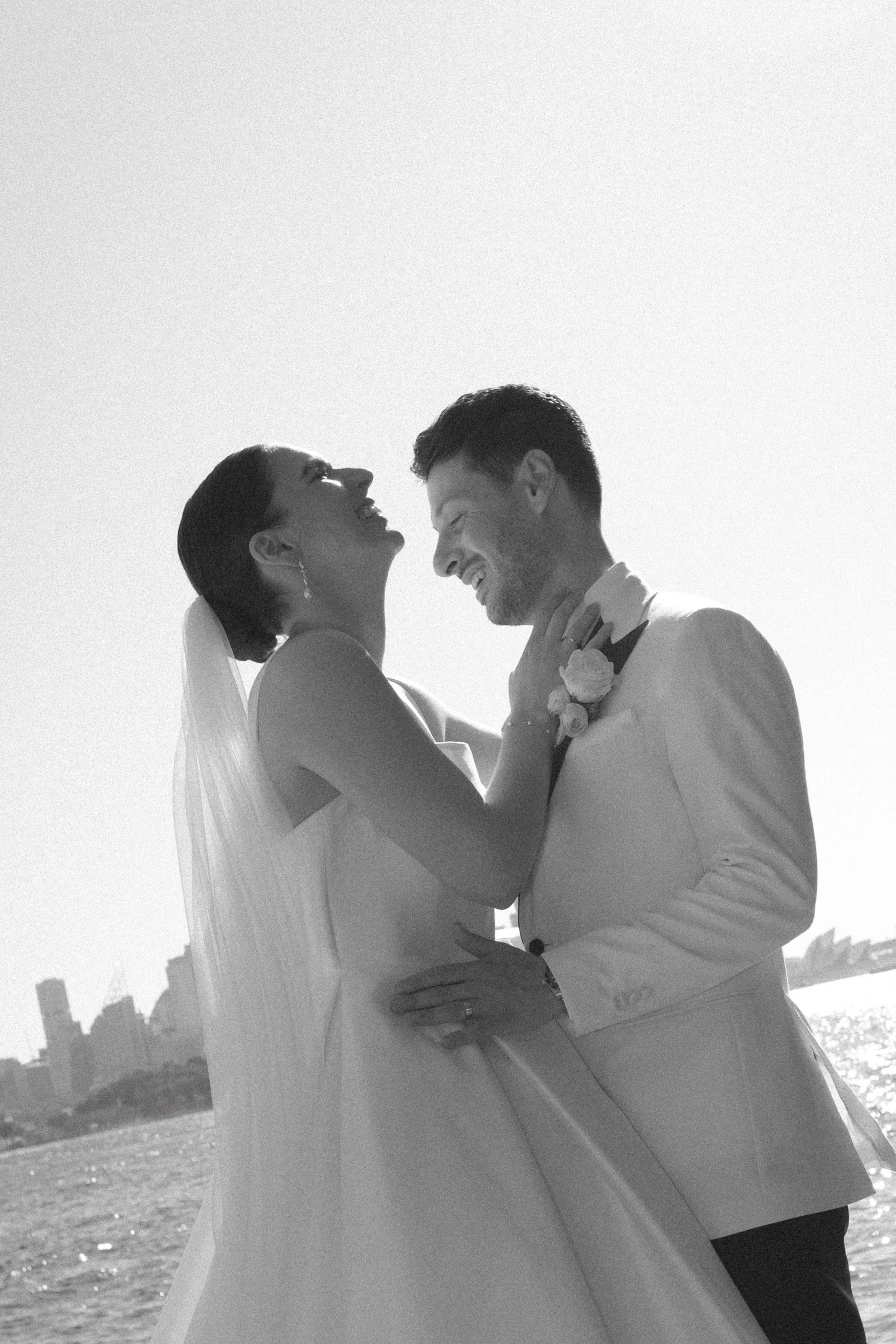 A black and white photo of a smiling bride and groom on their wedding day, holding each other close, with a city skyline and water in the background.
