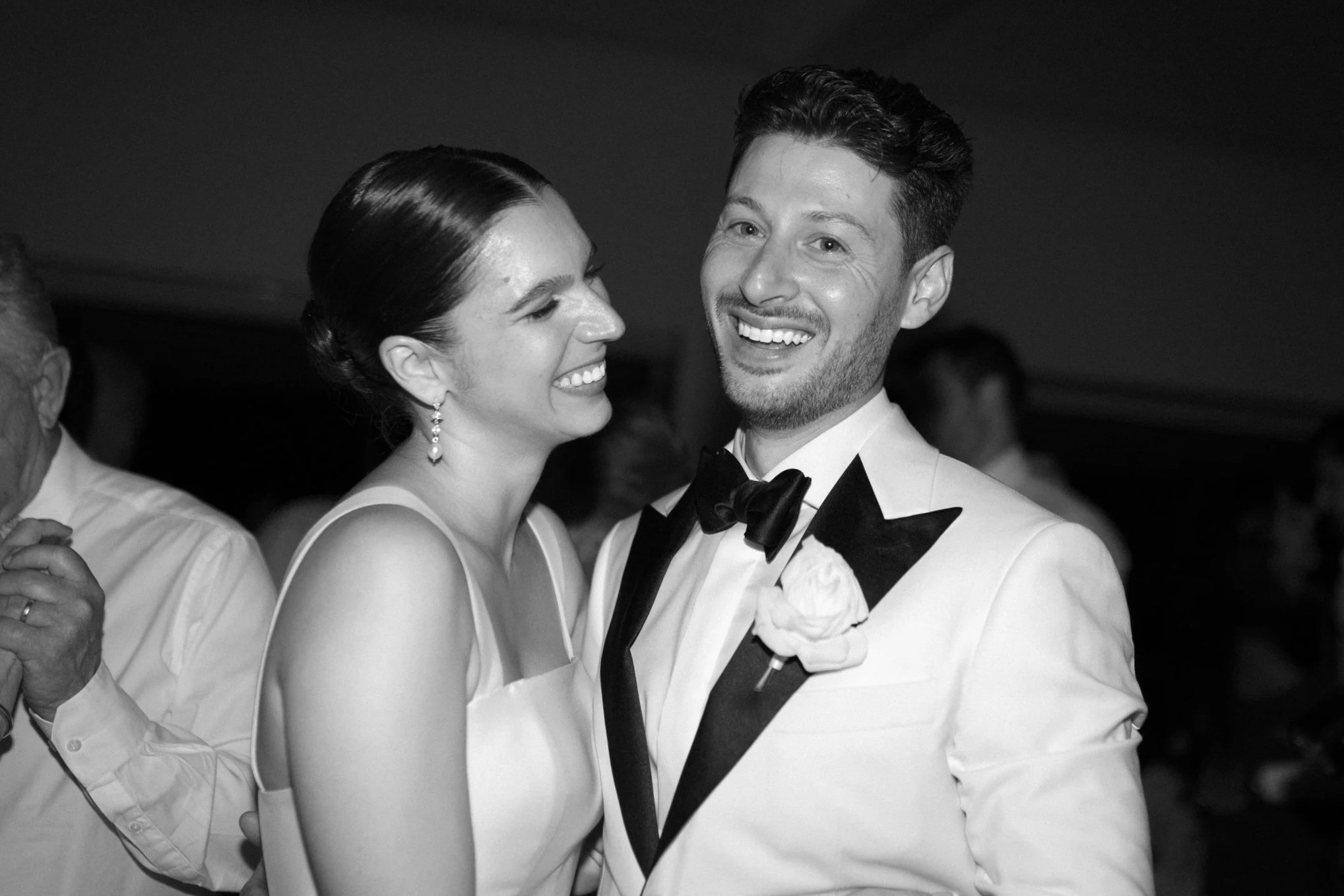 Black and white photo of a smiling couple at a formal event, the woman wearing a sleeveless dress and earrings, the man wearing a tuxedo with a bow tie and a boutonniere.