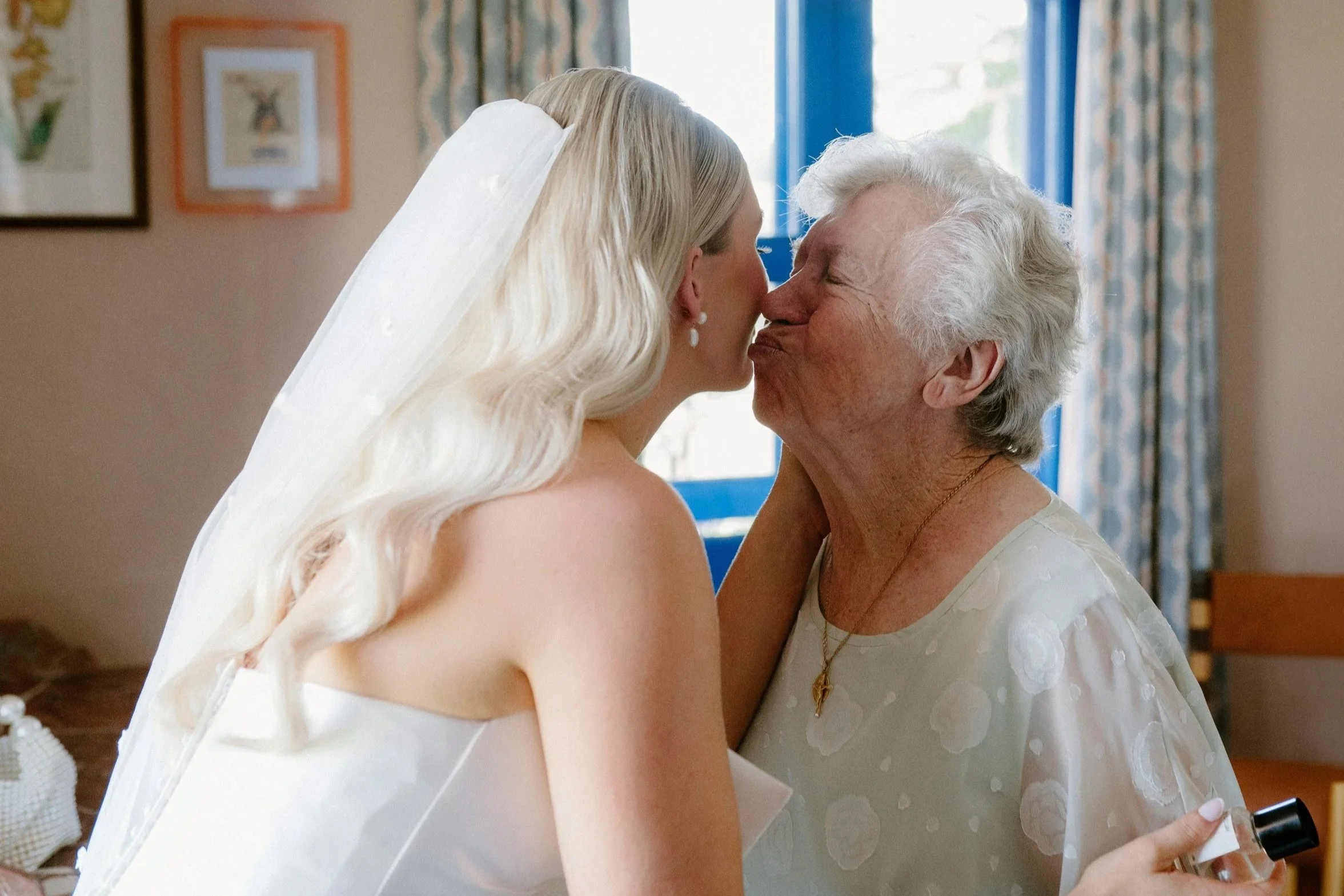 A bride in a white wedding dress sharing a kiss with an elderly woman, possibly her grandmother, in a room decorated with framed pictures and patterned curtains.