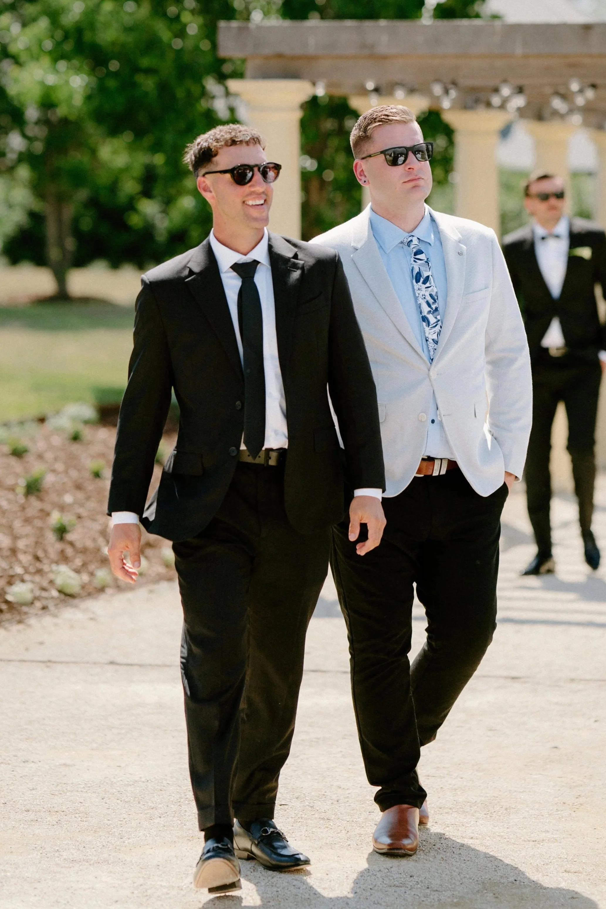 Two men in suits walking outdoors, one in a black suit and sunglasses, the other in a white blazer and floral tie, with a third man in the background also in formal attire.