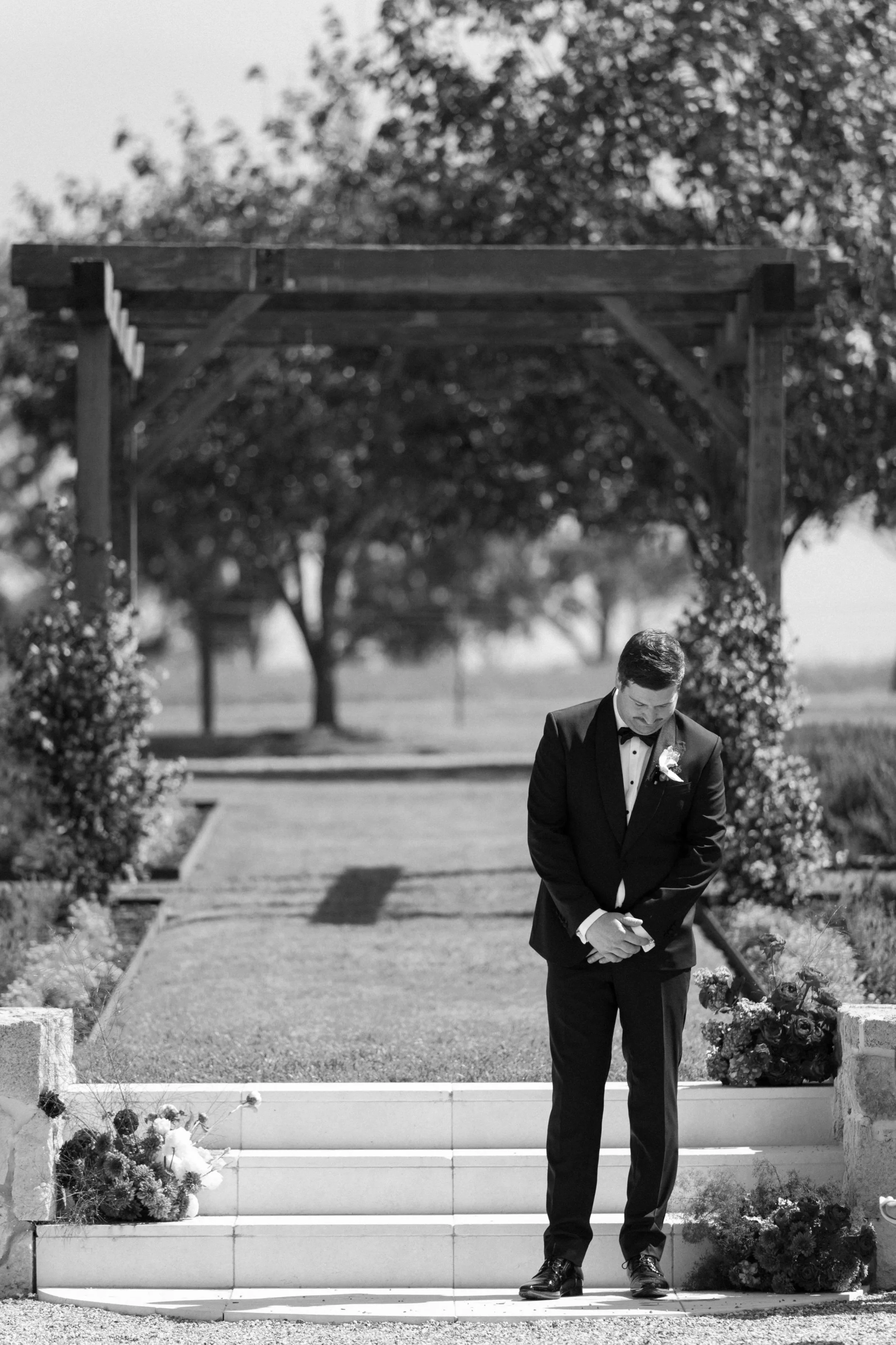 A man dressed in a tuxedo standing on outdoor stairs, bowing with head bowed and hands clasped, surrounded by flowers, with a garden and trees in the background.