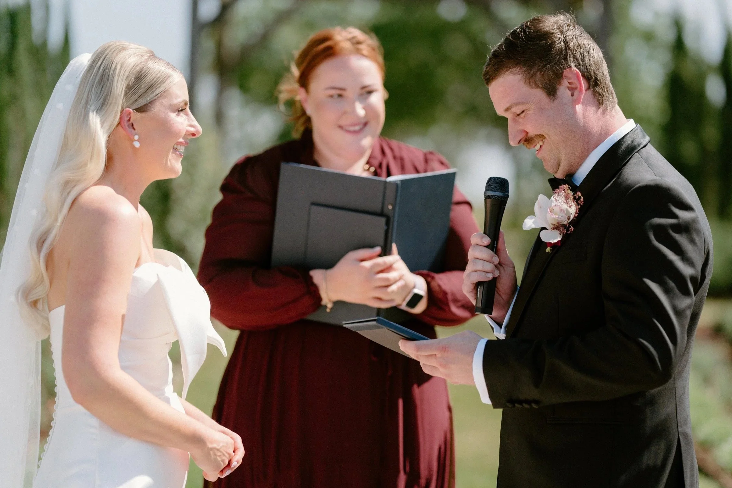 A bride and groom exchanging vows outdoors with an officiant holding a microphone and reading, all smiling and dressed formally.