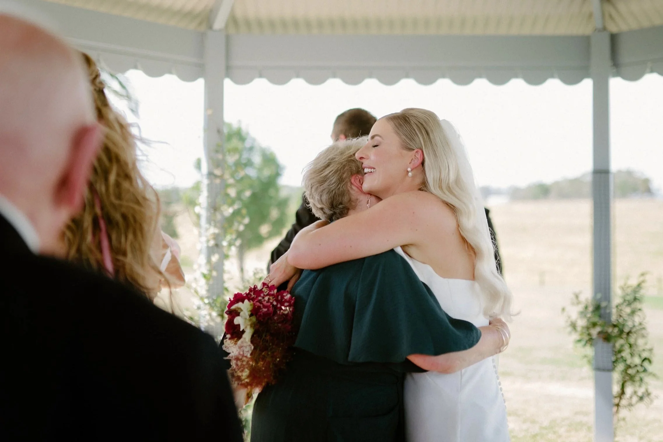 A woman in a white dress hugging an older woman in a dark dress at a wedding ceremony, with a blurred background and other people present.
