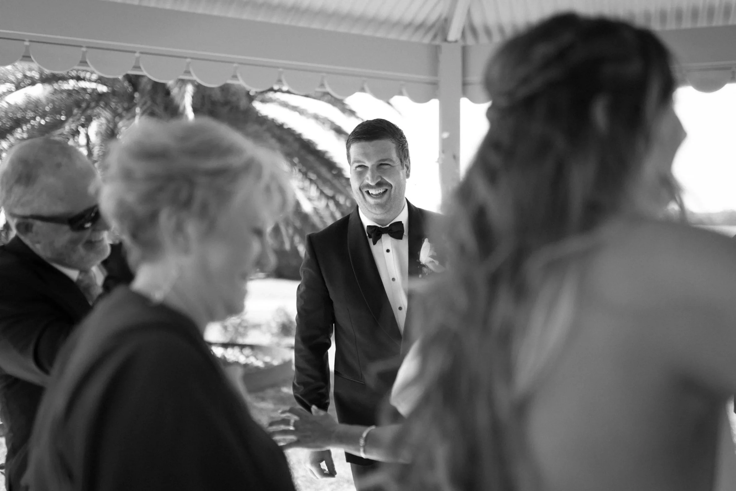 A groom in a tuxedo smiling at a wedding ceremony, surrounded by women, with some outdoors and a palm tree in the background.