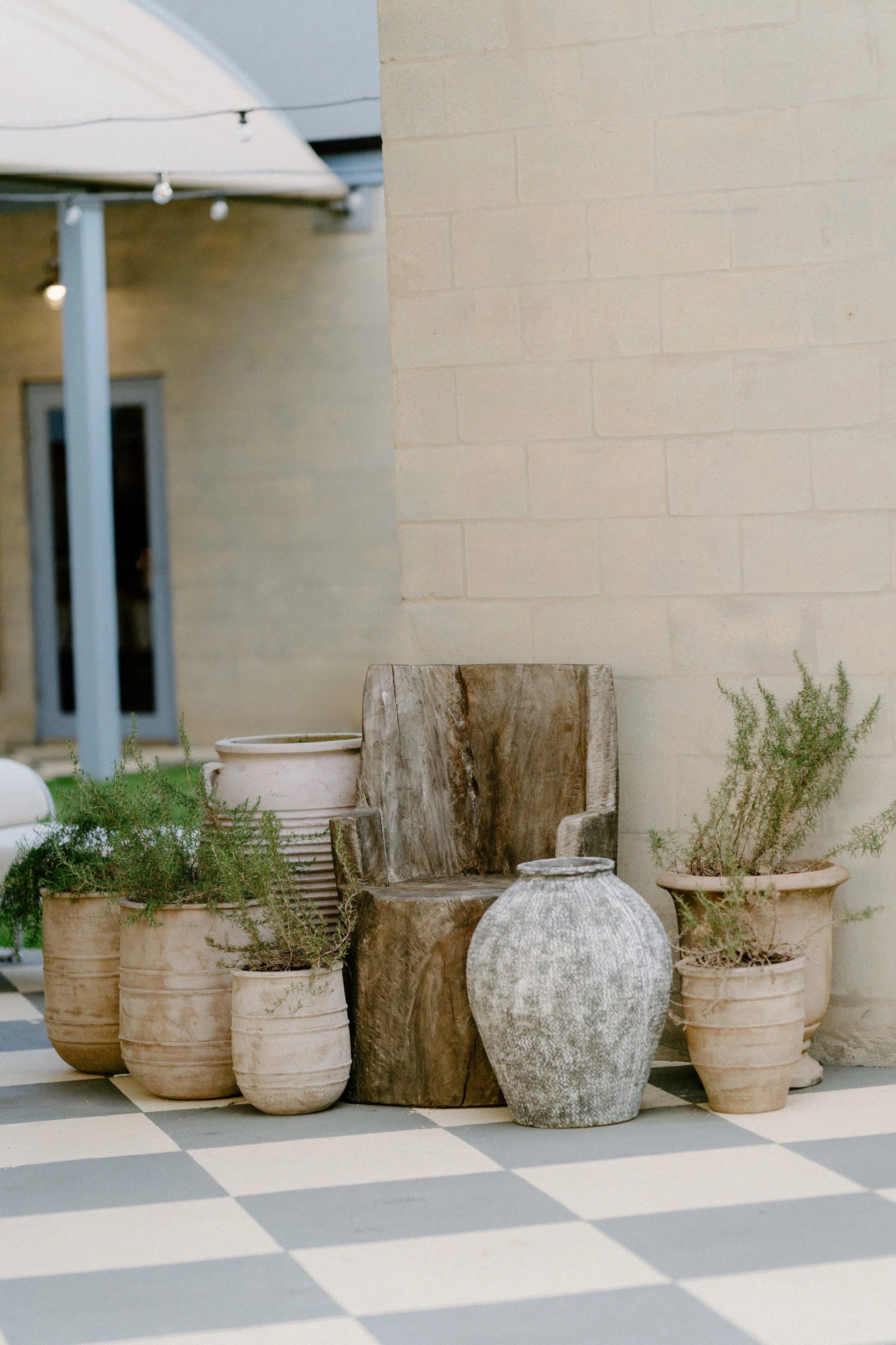 Clay pots and wooden chair outside on checkered floor with beige brick wall and building with string lights in background.