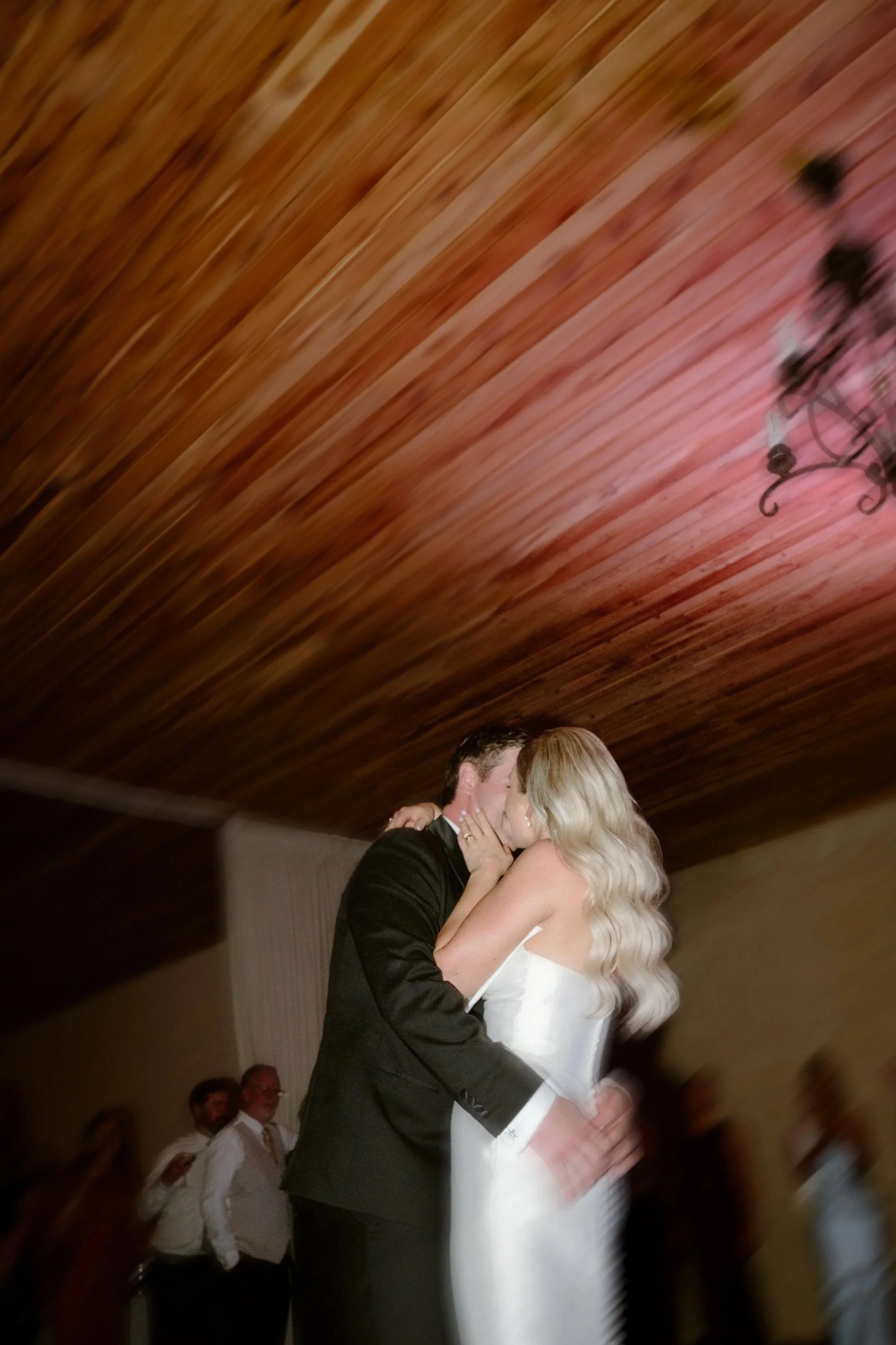 A bride and groom kiss during their wedding dance, with guests in the background and a wooden ceiling overhead.