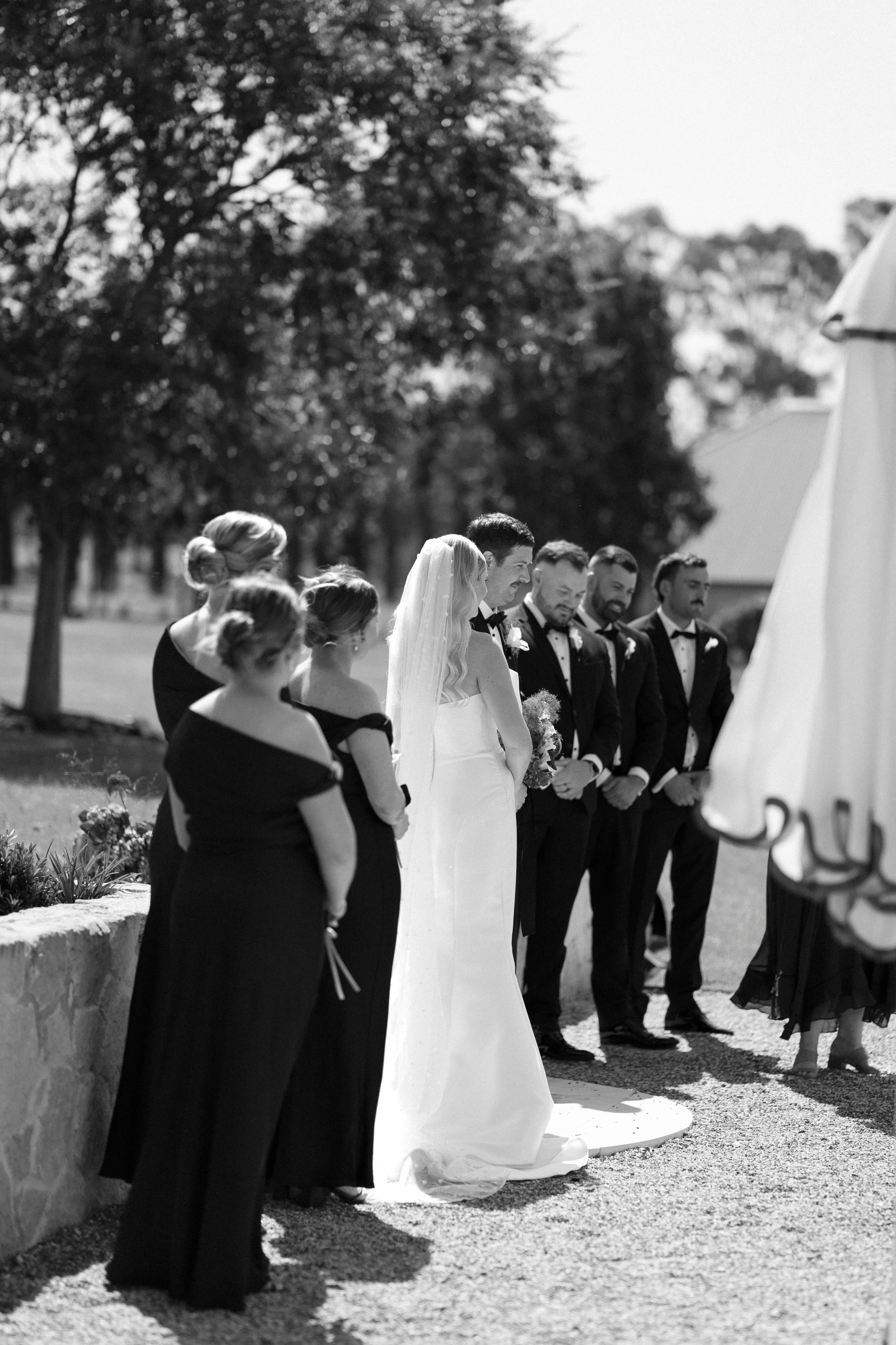 A group of people, including a bride in a wedding dress and veil, and several men in tuxedos, standing outdoors for a wedding ceremony.