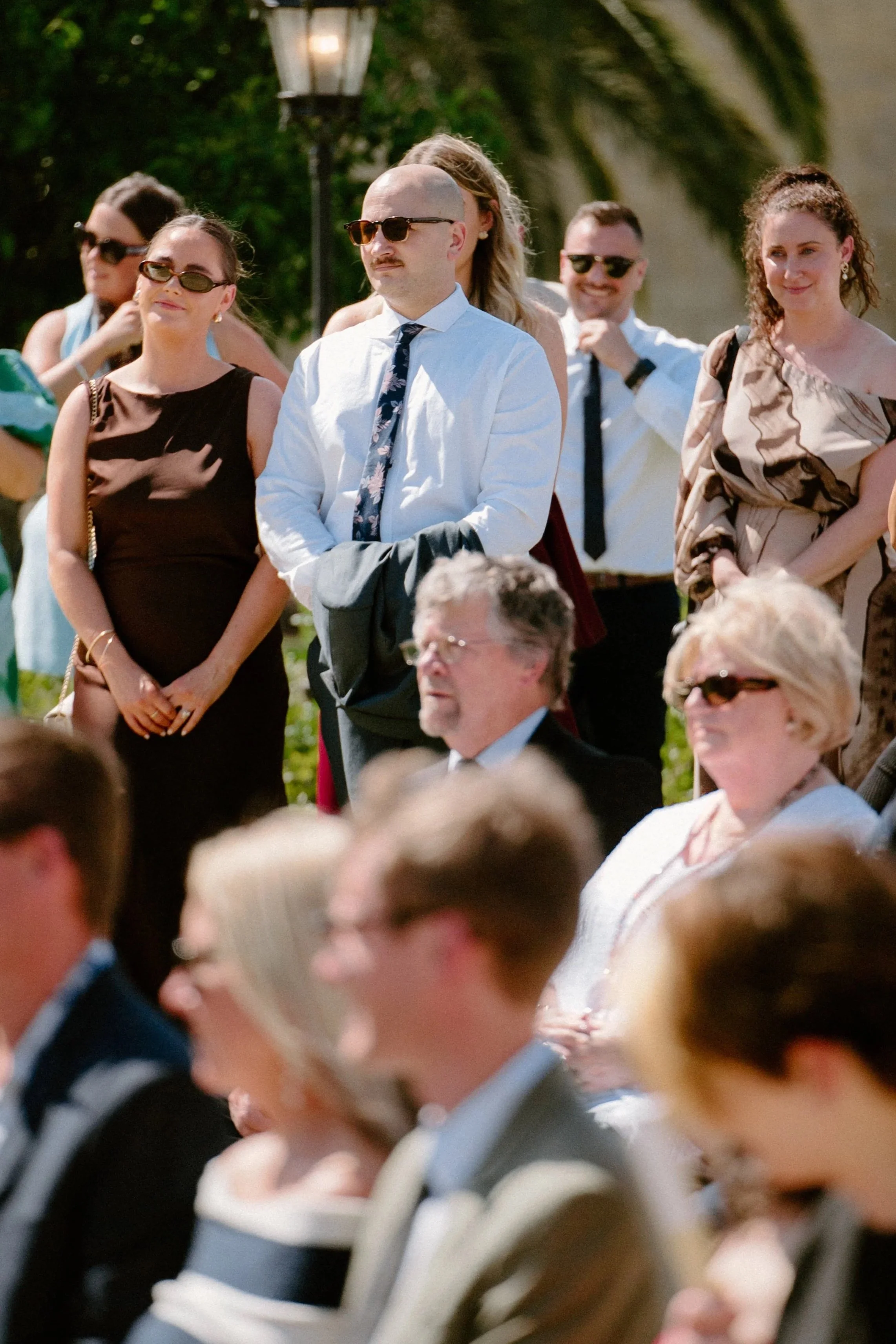 Group of people outdoors at a formal event or ceremony, with some wearing sunglasses, standing and sitting in front of a lamp post and greenery.