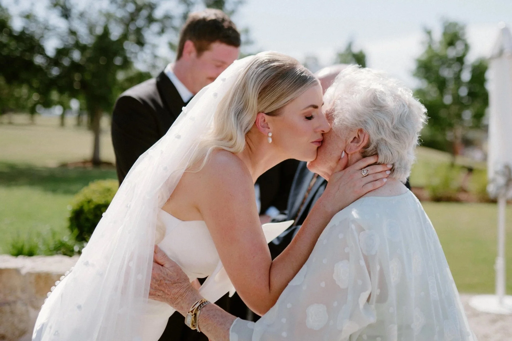 A bride kisses an elderly woman, likely her grandmother, during a wedding outdoors. The bride has blonde hair and is wearing a white dress and veil, while the elderly woman has white hair and is dressed in white. A man in a black suit stands behind t