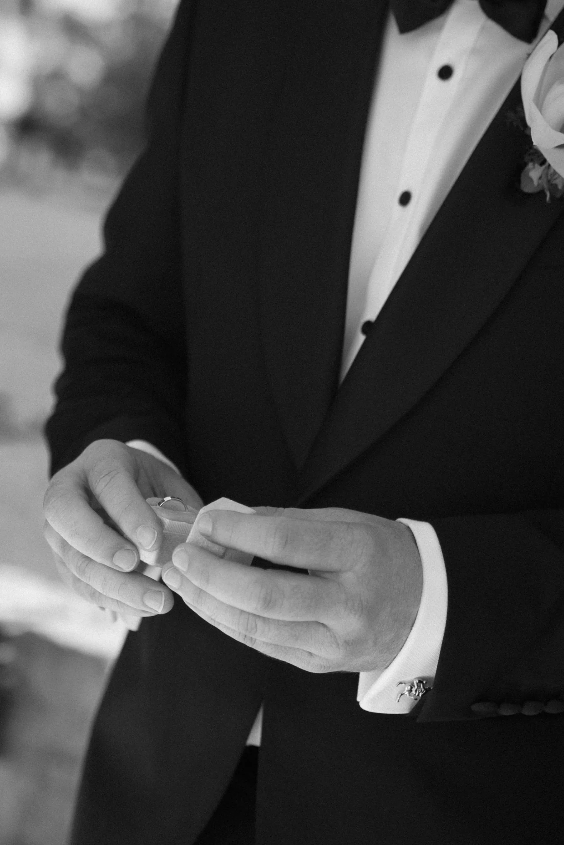 A groom in a tuxedo and bow tie holding a small object, possibly a ring box, with a blurred outdoor background.
